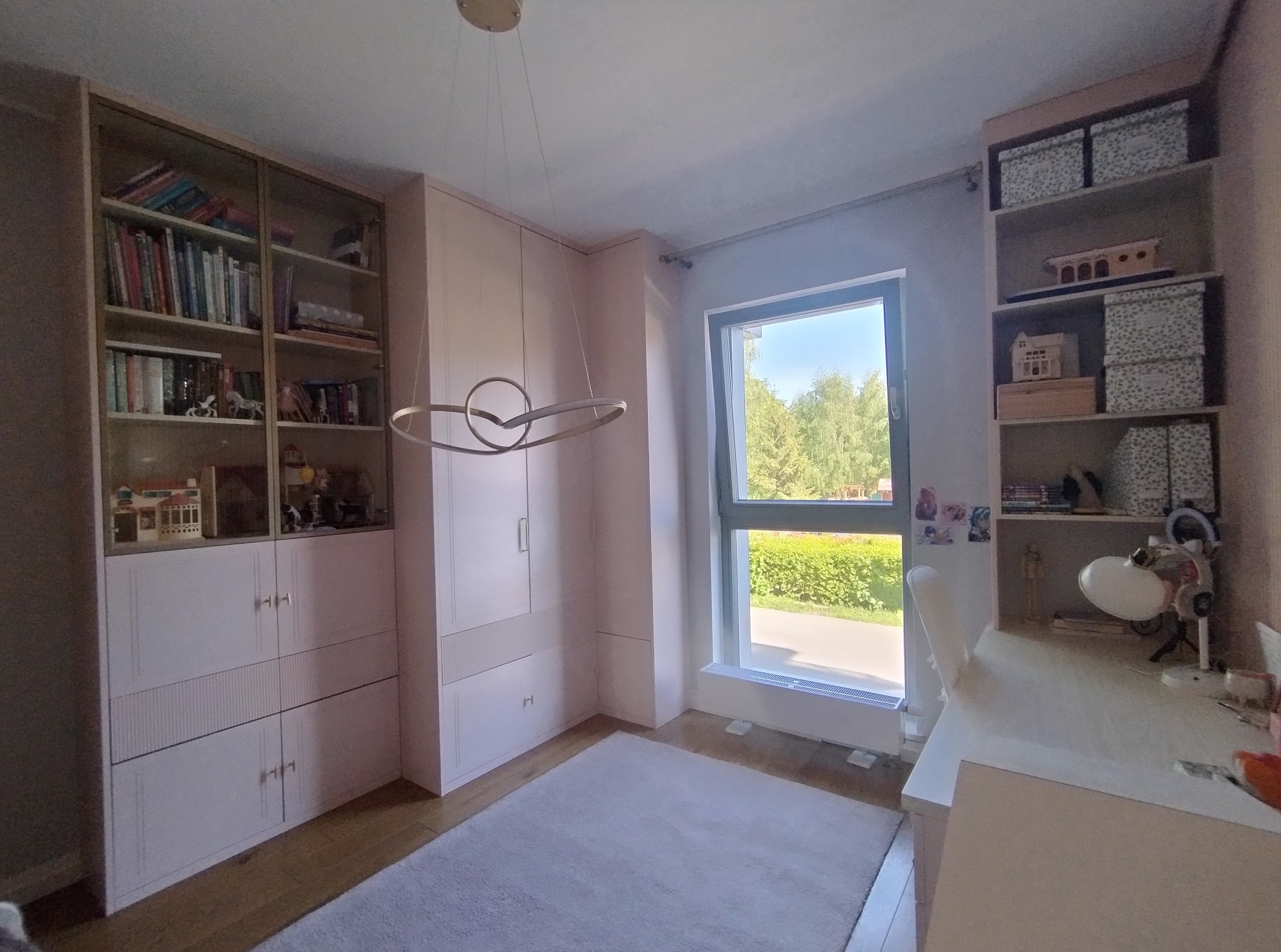 Jasny pokój z zabudową meblową: przeszklona biblioteczka, szafa z frezowanymi frontami i regał z dekoracjami. Okno z widokiem na zieleń. Całość w pastelowej kolorystyce.