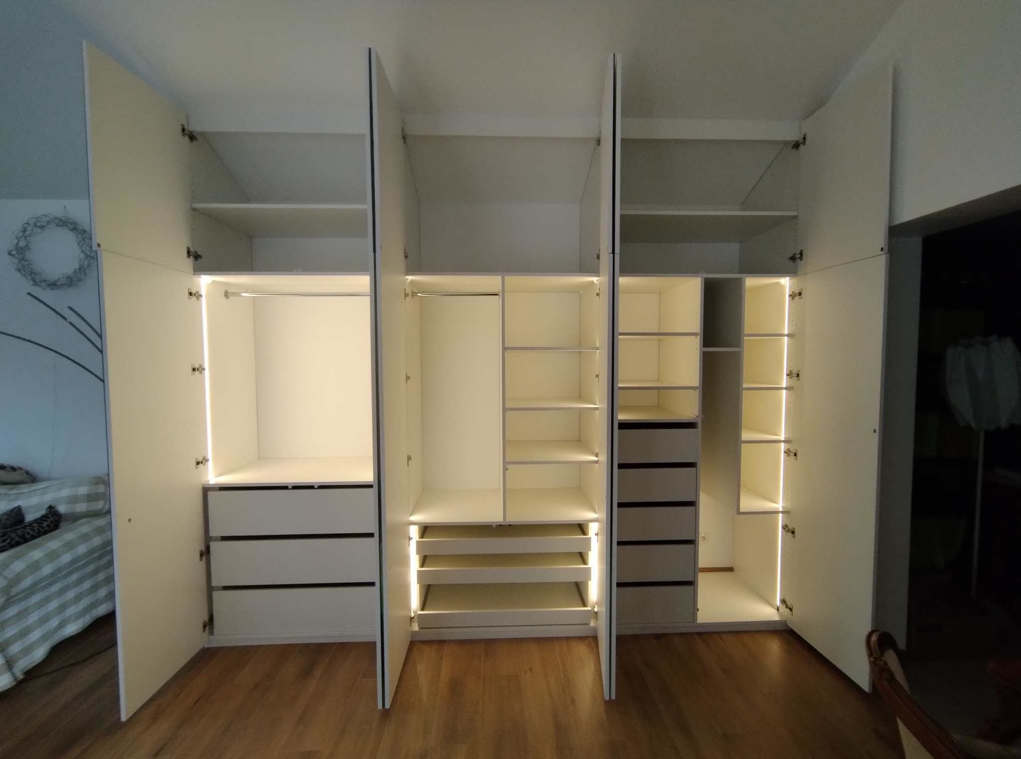 Otwarta, biała szafa wnękowa z oświetleniem LED, ukazująca półki, szuflady i drążki na ubrania. Widoczny parkiet na podłodze i fragment sypialni w tle.
