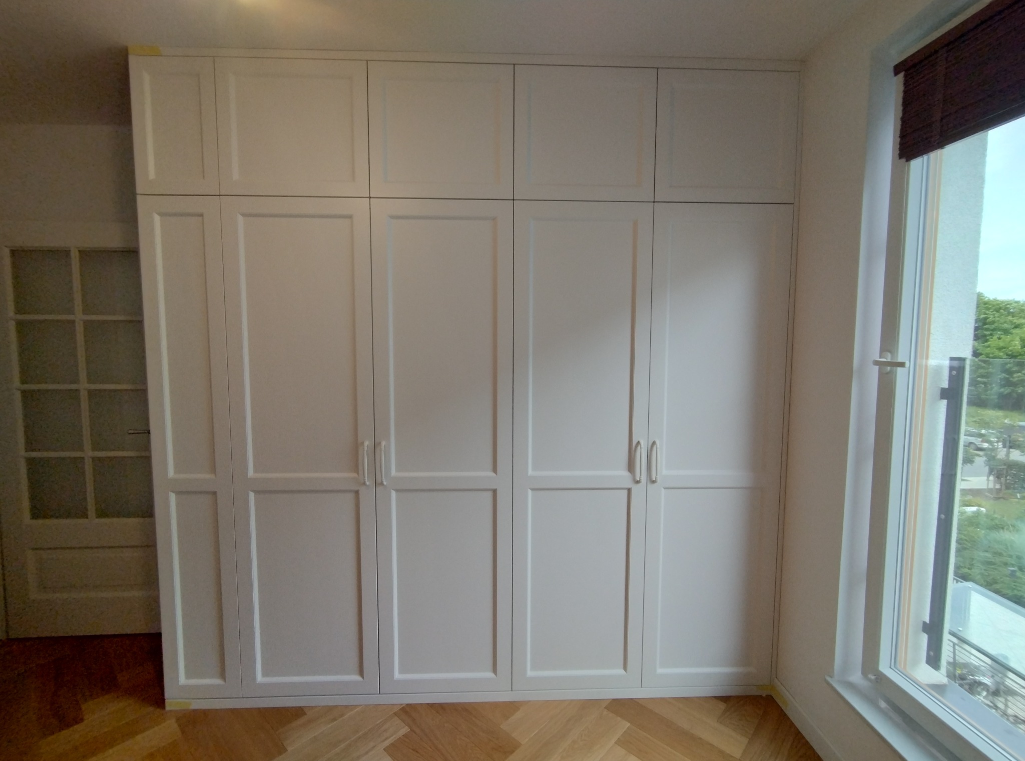 Biała szafa wnękowa z czterema drzwiami i górnymi szafkami, w jasnym pomieszczeniu z parkietem i oknem z widokiem na zieleń. Klasyczny design, proste uchwyty.