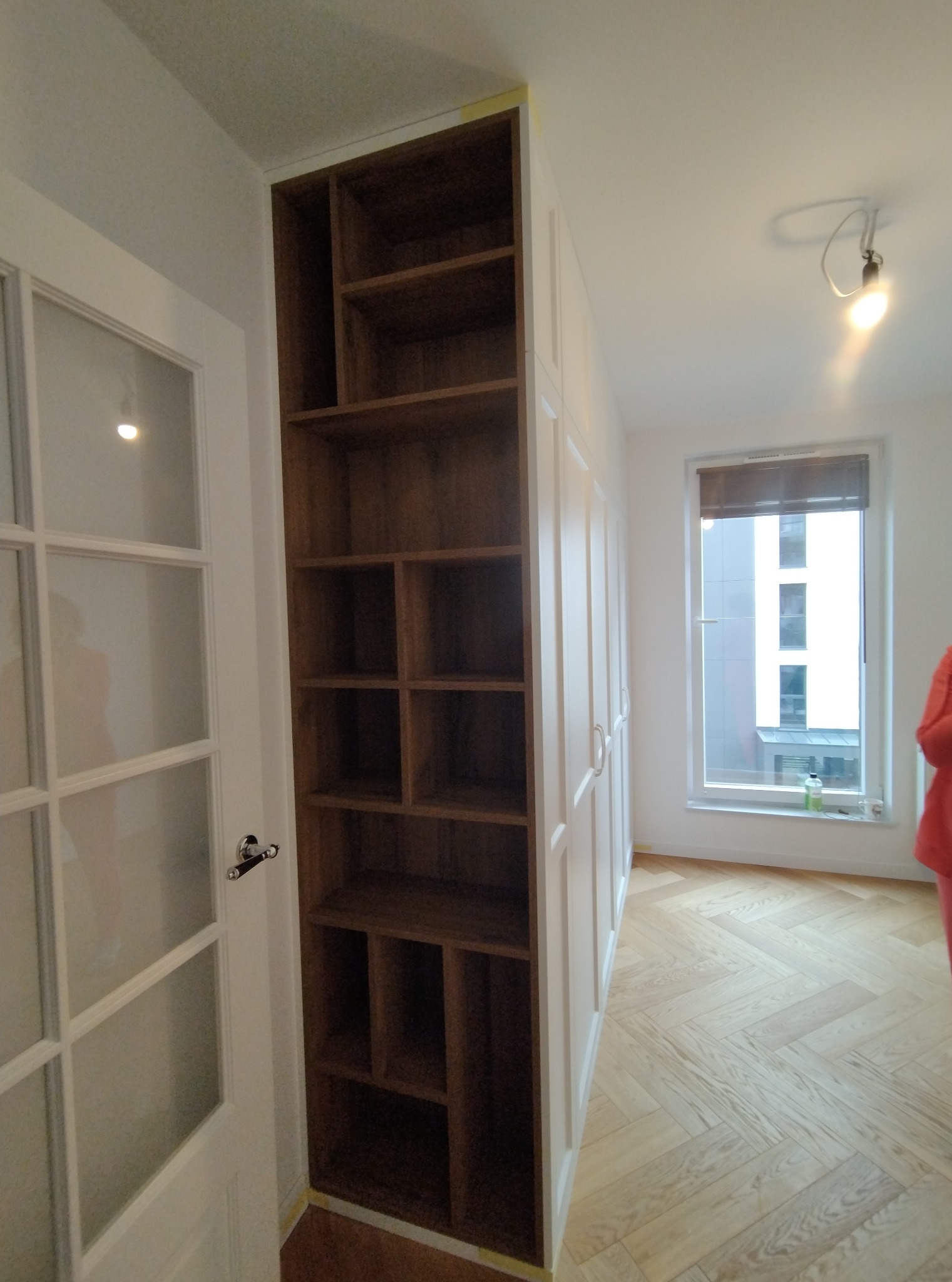 Wnętrze z drewnianą szafą wnękową z otwartymi półkami, przylegającą do białej szafy zamykanej, na parkiecie w jodełkę, z widokiem na okno.