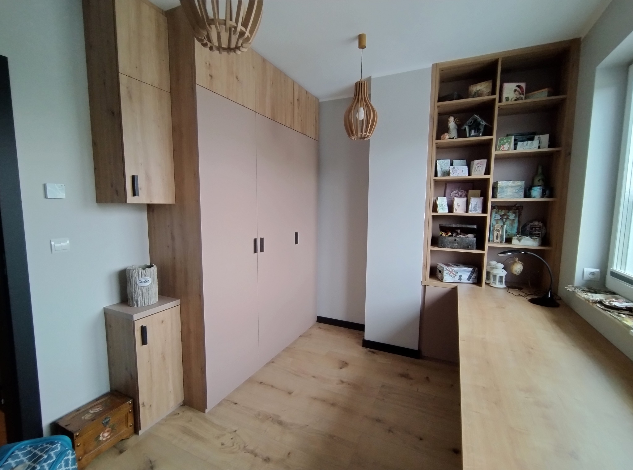 Jasne wnętrze z zabudową: szafa z różowymi frontami i drewnianymi górnymi szafkami, regał z dekoracjami i drewniany blat wzdłuż okna. Drewniana podłoga i lampy.