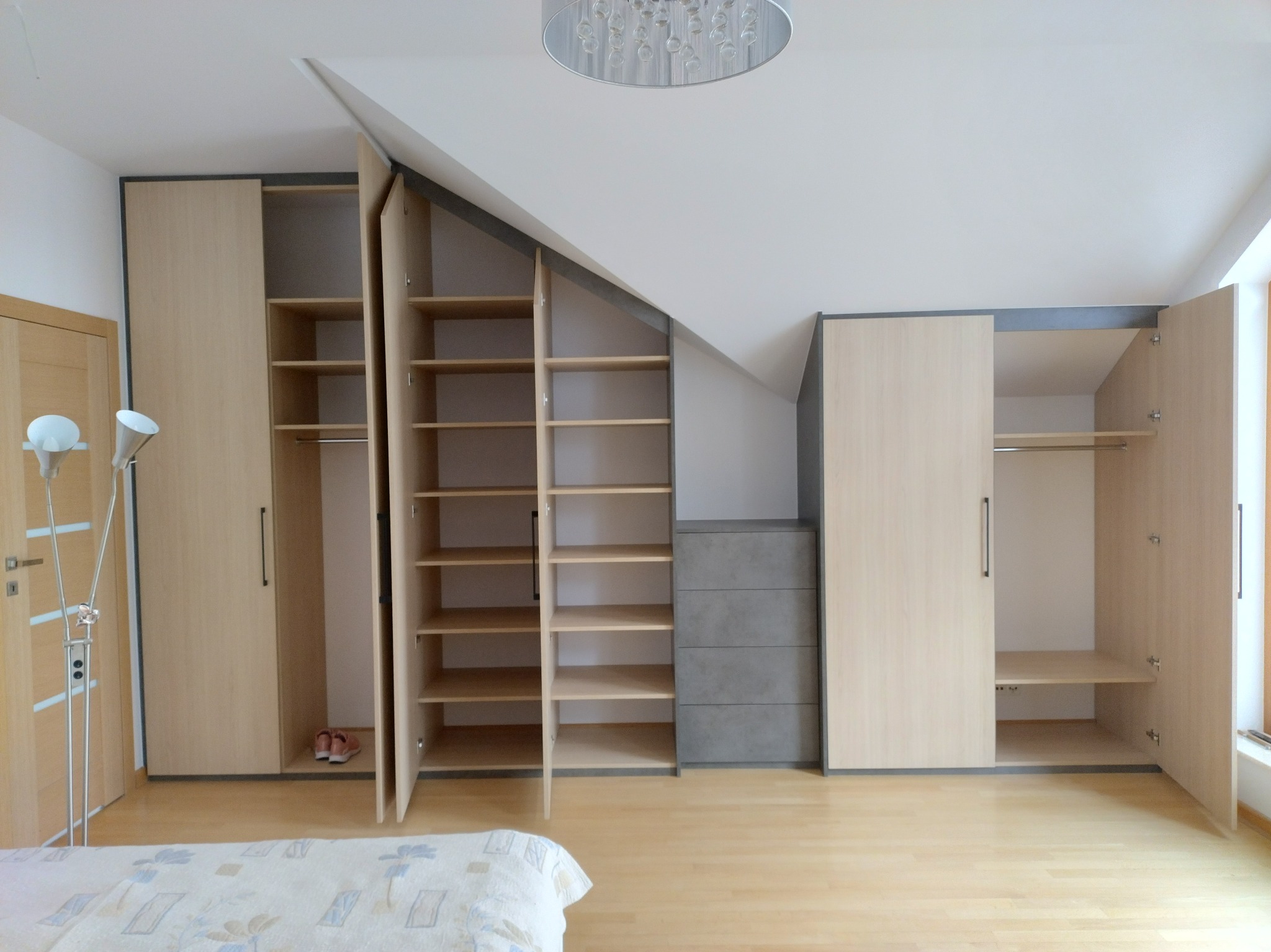 Jasna szafa wnękowa z półkami i szufladami, dopasowana do skosu dachu. Częściowo otwarte drzwi ukazują wnętrze. Widoczna podłoga z paneli i fragment łóżka.