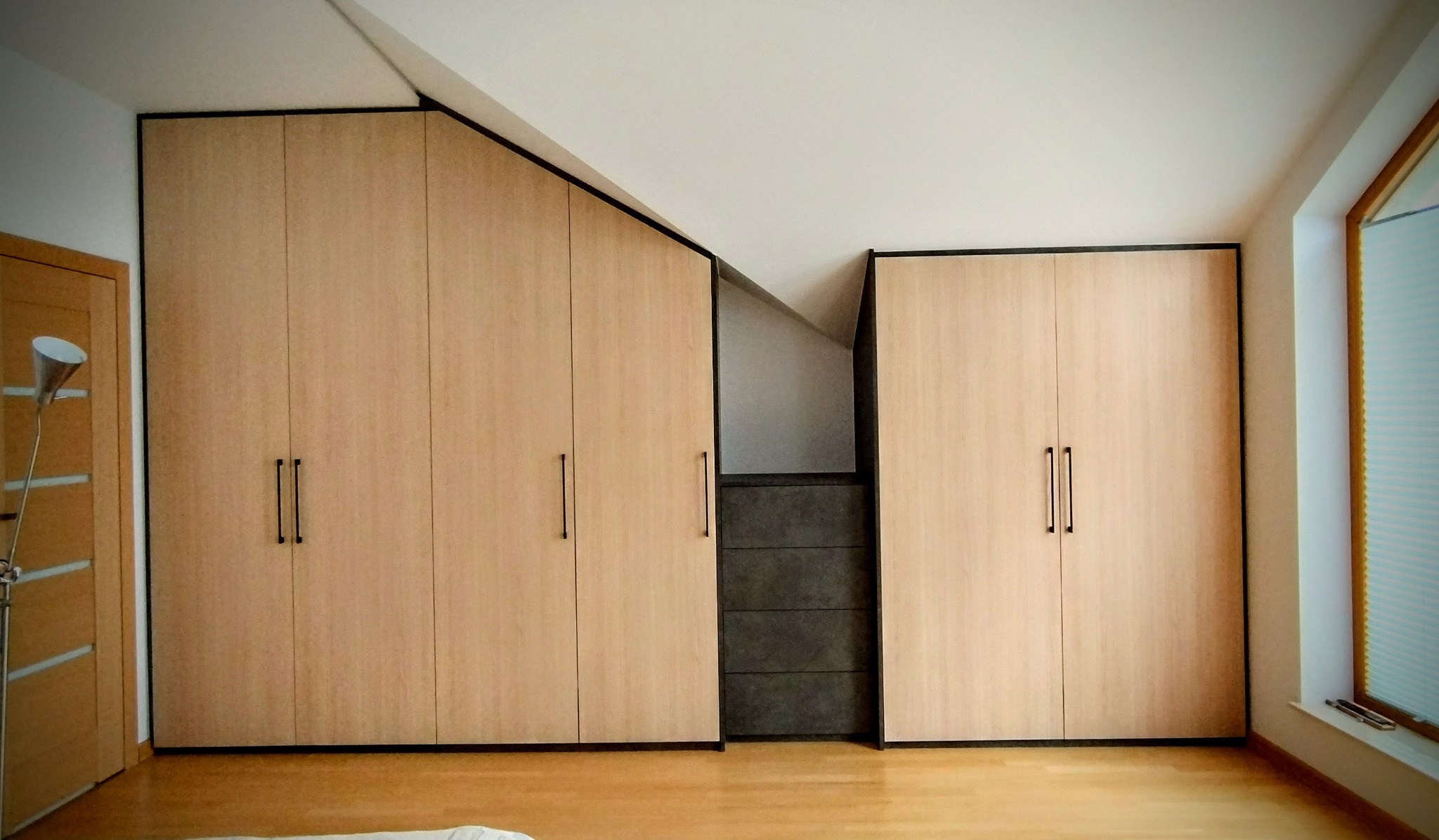 Jasne, drewniane szafy wnękowe z czarnymi obramowaniami i uchwytami, dopasowane do skosu dachu. Widoczna podłoga z paneli i okno z roletą.