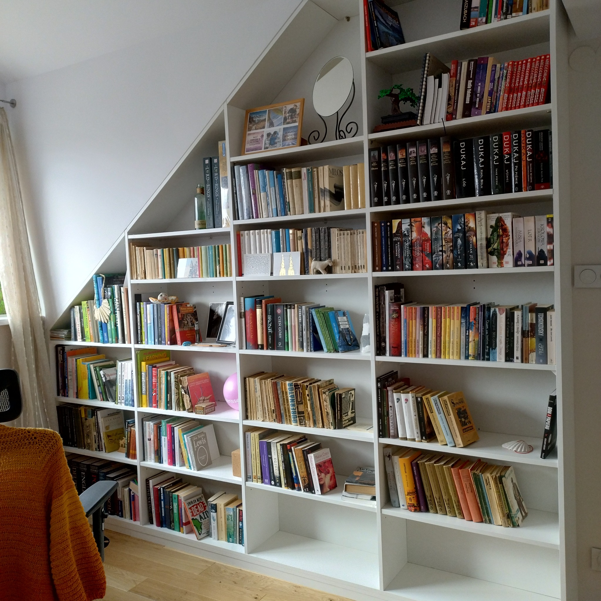 Biała, zabudowana biblioteczka na książki pod skosem dachu, wypełniona książkami, dekoracjami i pamiątkami. Widoczne drewniane półki i różnorodna kolorystyka okładek.