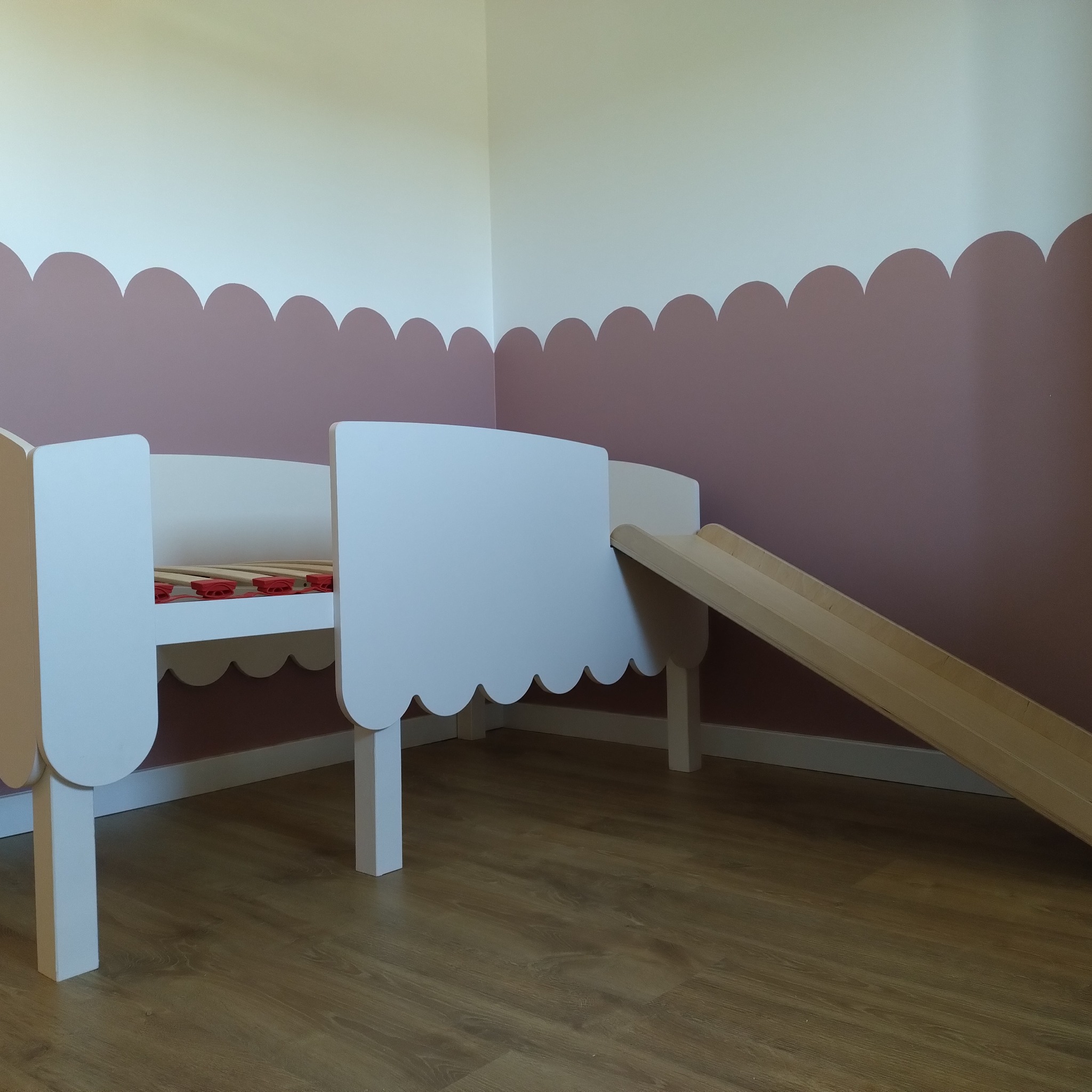 Białe łóżko dziecięce z drewna ze zjeżdżalnią, na podłodze panele, ściana w pastelowych kolorach. Nietypowy kształt ramy łóżka.