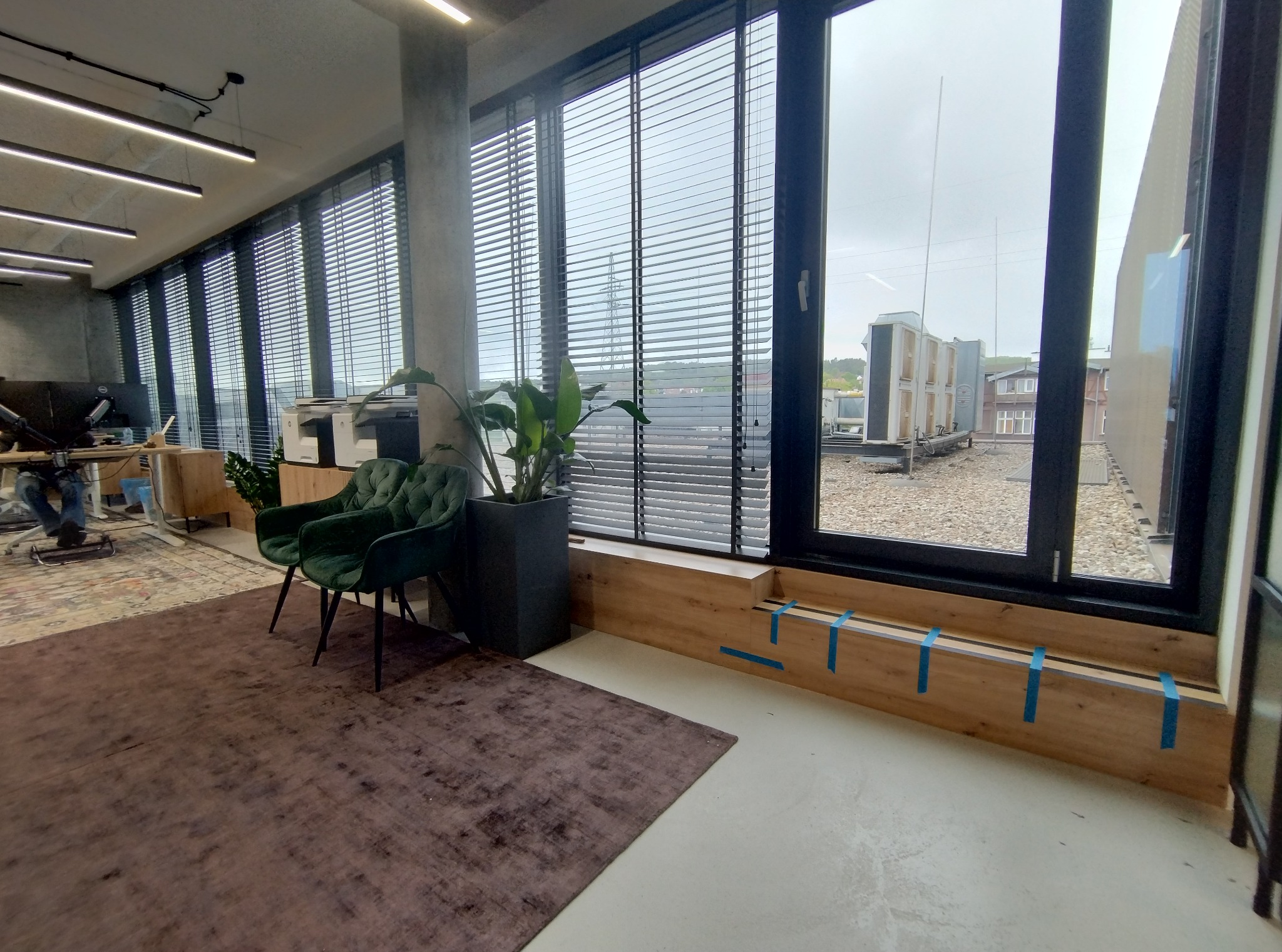 Nowoczesne wnętrze biurowe z drewnianą zabudową okienną zabezpieczoną taśmą malarską, zielonymi krzesłami i dywanem. Widok na zewnątrz przez żaluzje.