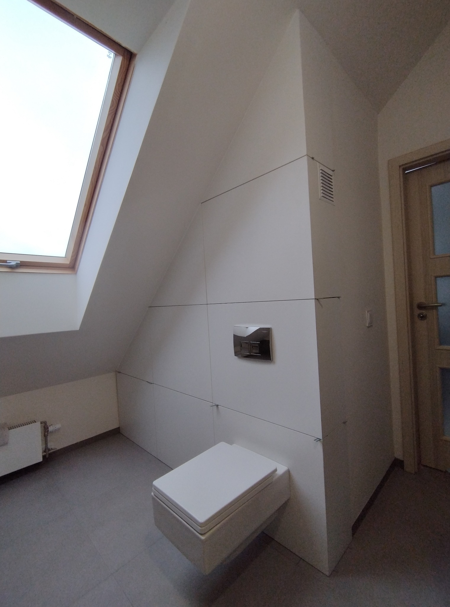 Biała zabudowa wnęki w łazience na poddaszu ze spłuczką podtynkową i wiszącą toaletą. Widoczne okno dachowe i grzejnik.
