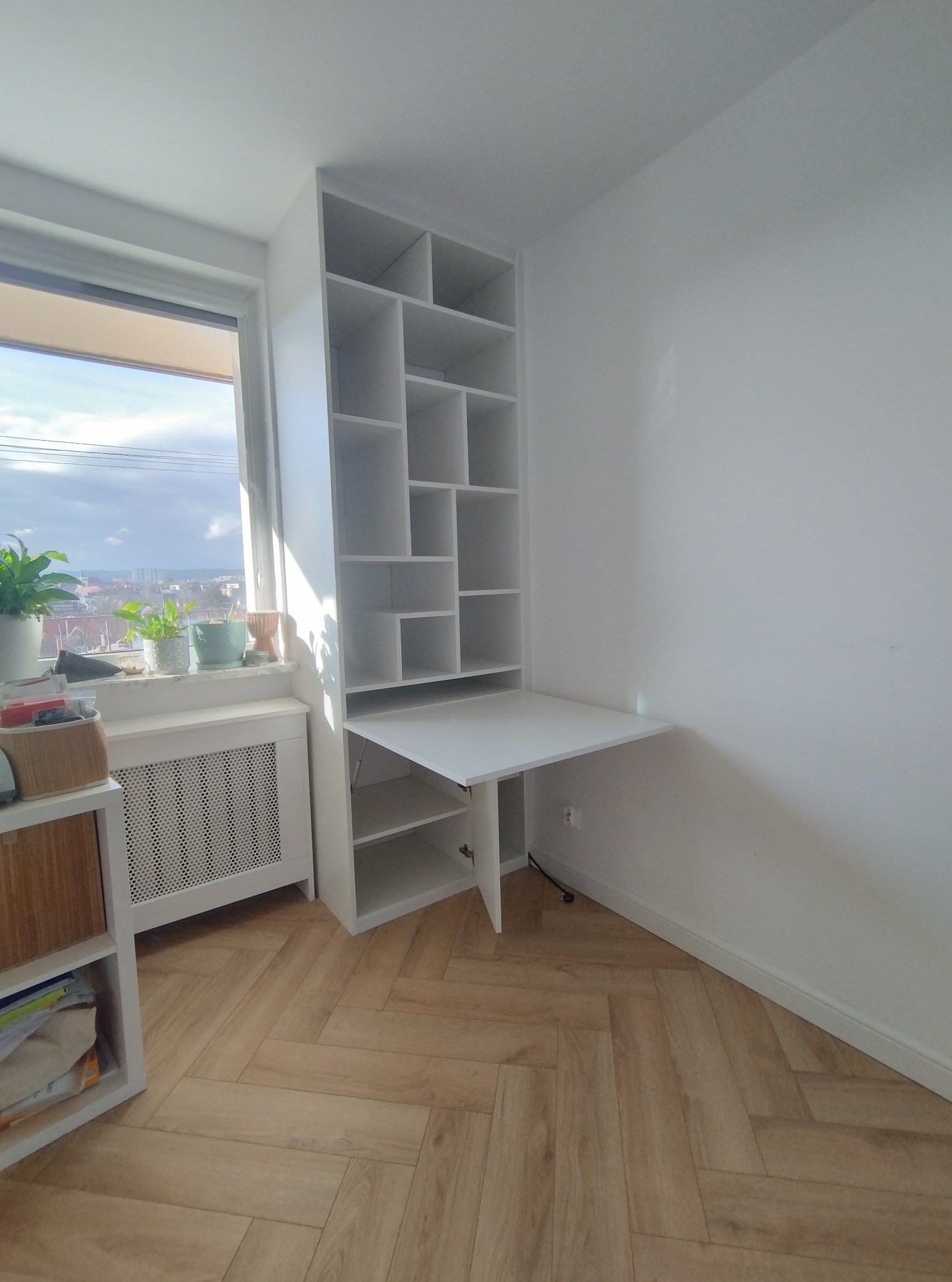 Biała szafa wnękowa z rozkładanym blatem biurka, umieszczona przy oknie z widokiem na miasto. Jasne wnętrze, parkiet w jodełkę. Minimalistyczny design.