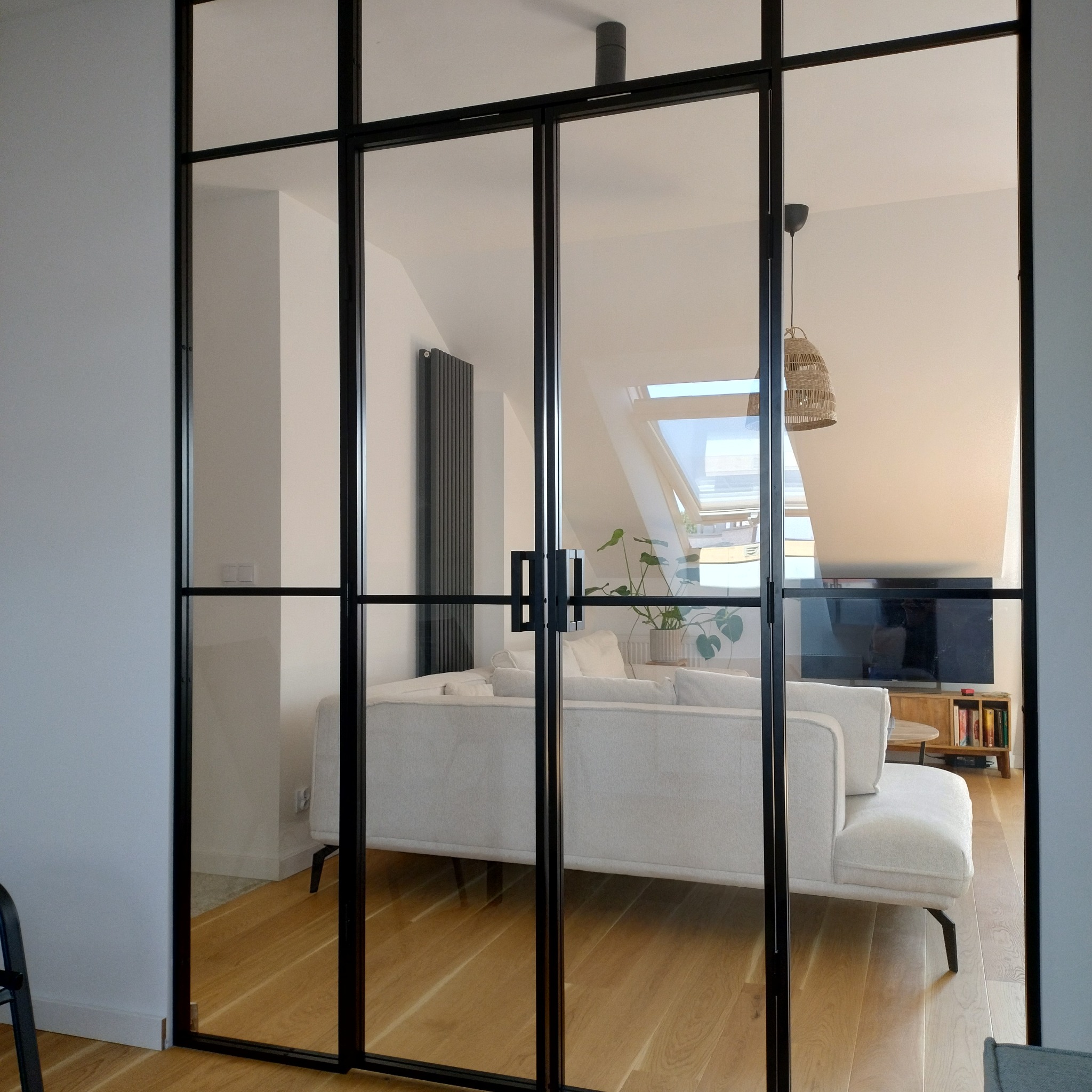 Loftowe drzwi przesuwne z czarnymi szprosami, oddzielające pokój z jasną sofą i drewnianą podłogą. Widoczny grzejnik i skos dachu z oknem.