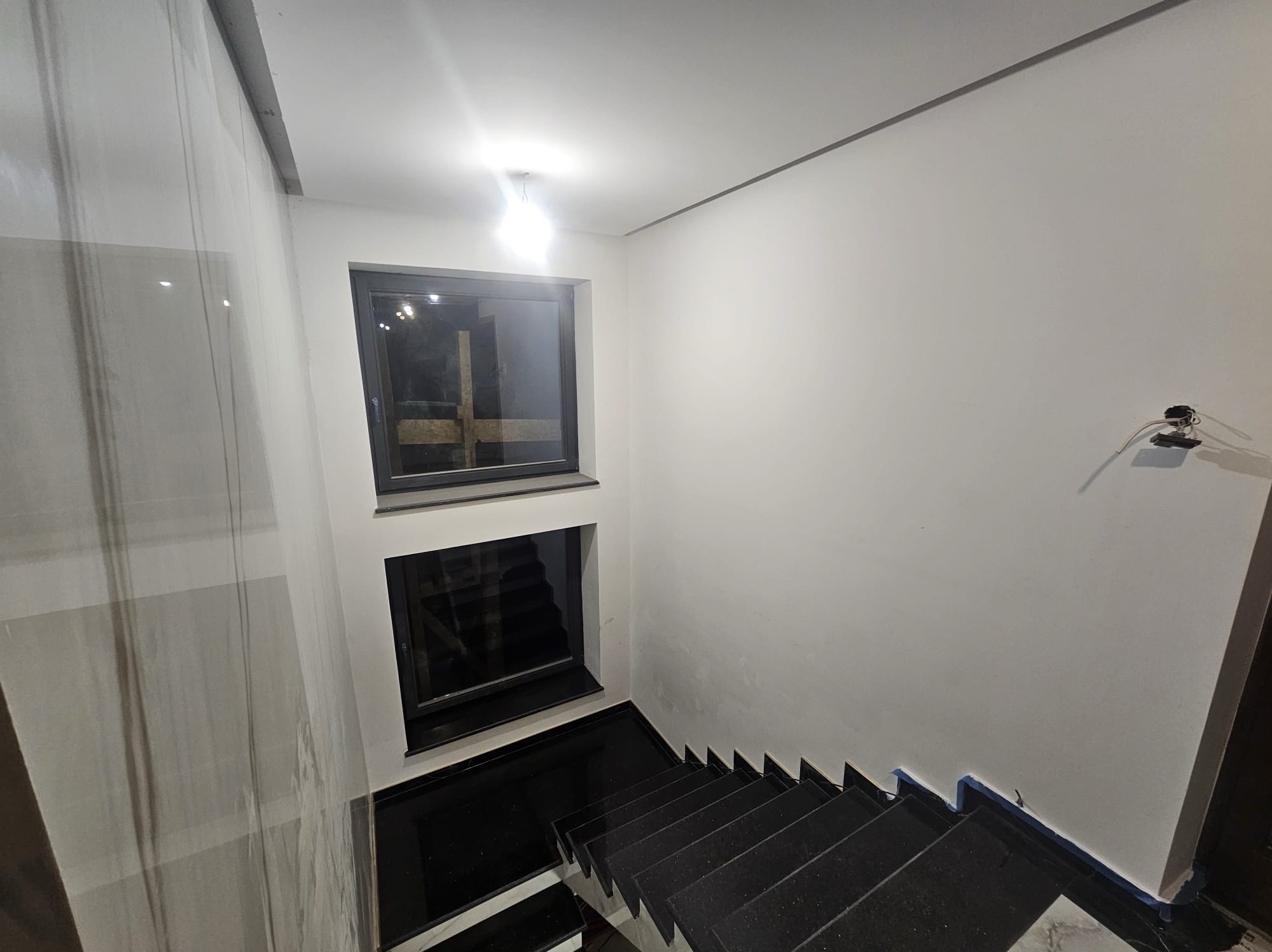 Nowoczesne schody z czarnego granitu, dwa okna w czarnych ramach, białe ściany. Minimalistyczne wykończenie wnętrza, widok z góry na dół.