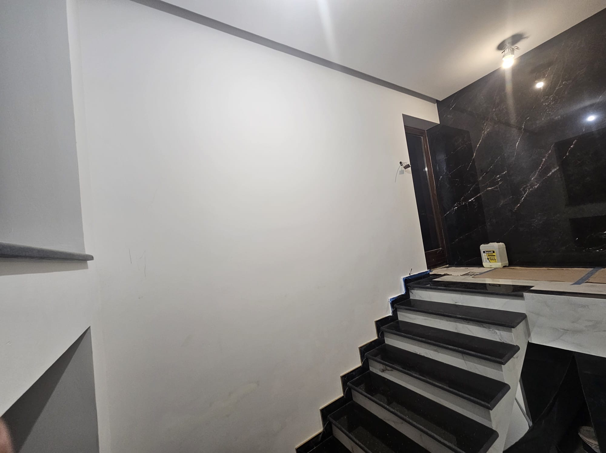 Wykończone schody z czarnego kamienia prowadzące na górę, kontrastujące z białą ścianą i marmurową okładziną. Widoczny fragment sufitu z oświetleniem.