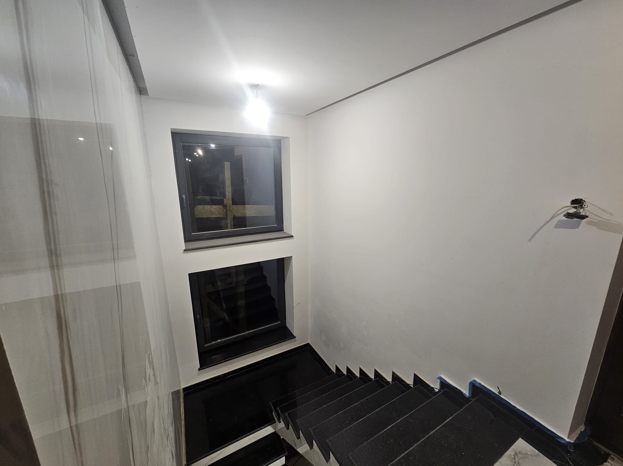 Wnętrze klatki schodowej z czarnymi schodami i dwoma oknami. Ściany białe, częściowo wykończone. Widoczne zabezpieczenia malarskie.