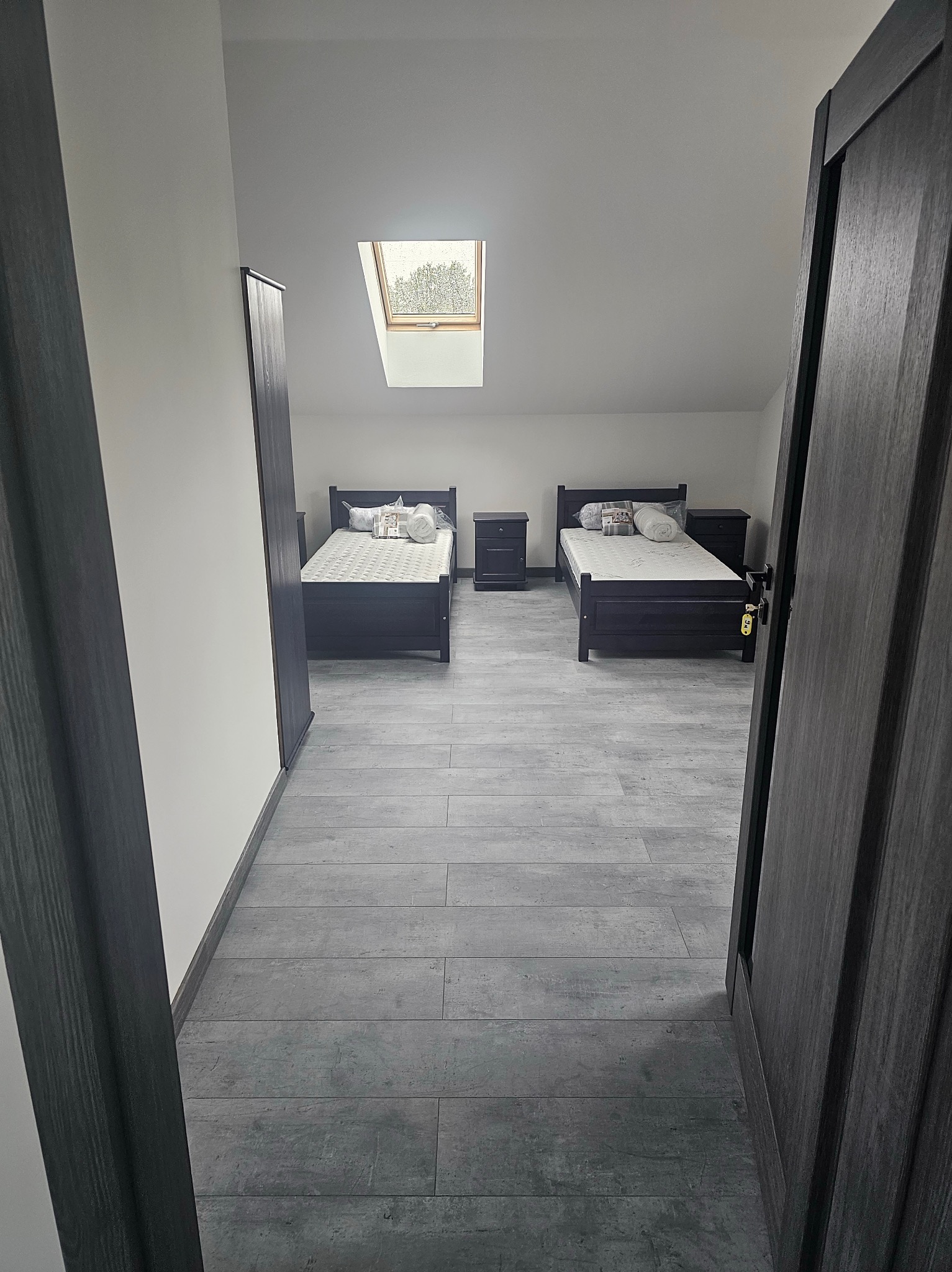 Pokój na poddaszu z dwoma łóżkami i oknem dachowym. Szare panele podłogowe, ciemne meble. Widok zza otwartych drzwi, z perspektywy wejścia do pomieszczenia.