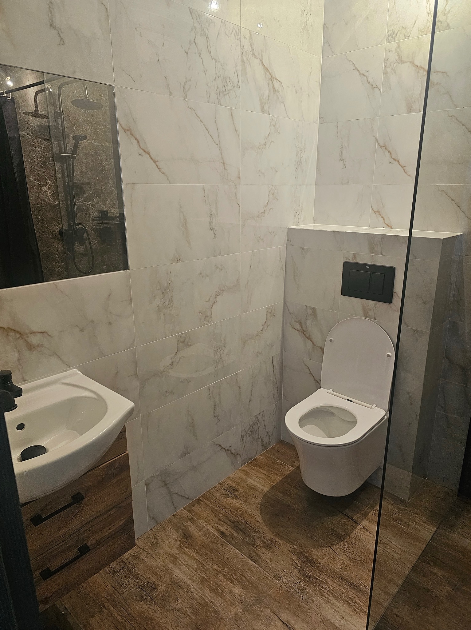 Nowoczesna łazienka z marmurowymi płytkami, drewnopodobną podłogą i minimalistyczną armaturą. Umywalka z szafką i toaleta w zabudowie.