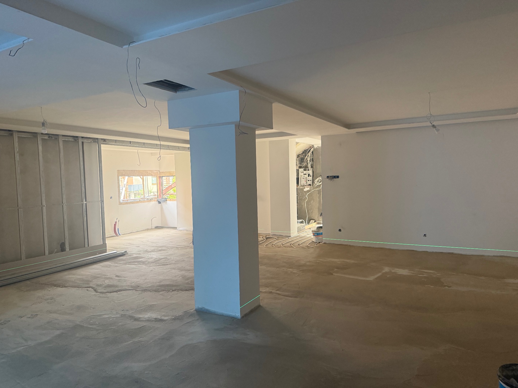 Wnętrze w trakcie prac wykończeniowych: surowy beton na podłodze, ściany przygotowane do malowania, widoczne instalacje elektryczne i rury ogrzewania podłogowego.