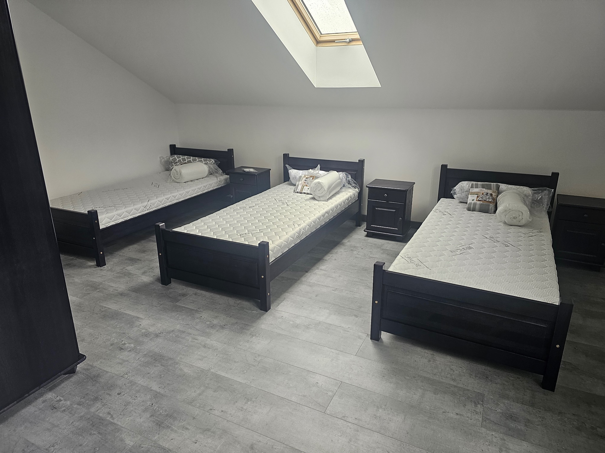 Pokój na poddaszu z trzema łóżkami z czarnymi ramami i szafkami nocnymi. Okno dachowe wpuszcza światło. Podłoga wyłożona panelami w odcieniach szarości.