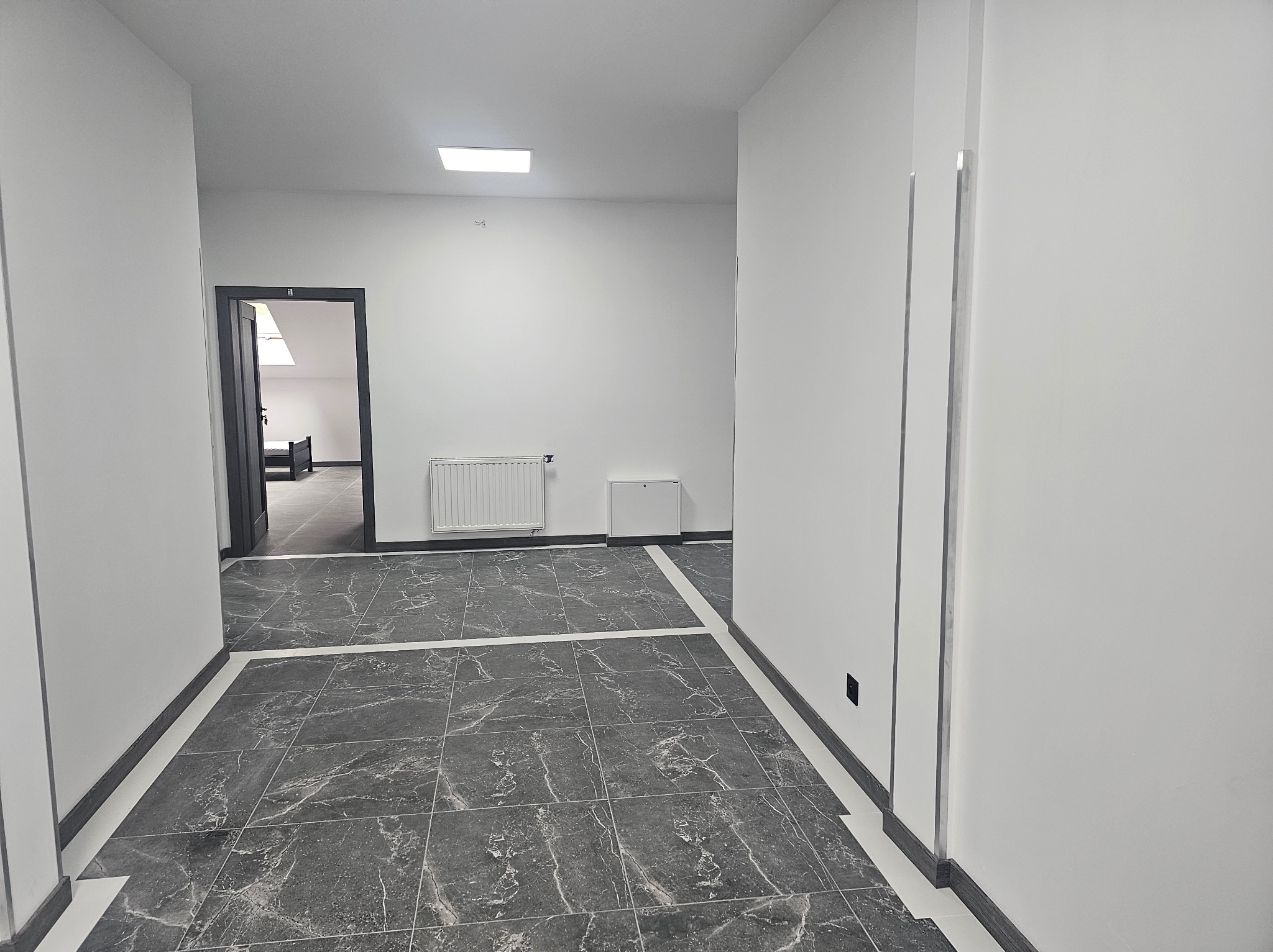 Korytarz z szarą marmurową podłogą i białymi ścianami. Widoczne drzwi do pokoju z łóżkiem i grzejnik na ścianie. Minimalistyczny design, nowoczesne wykończenie.