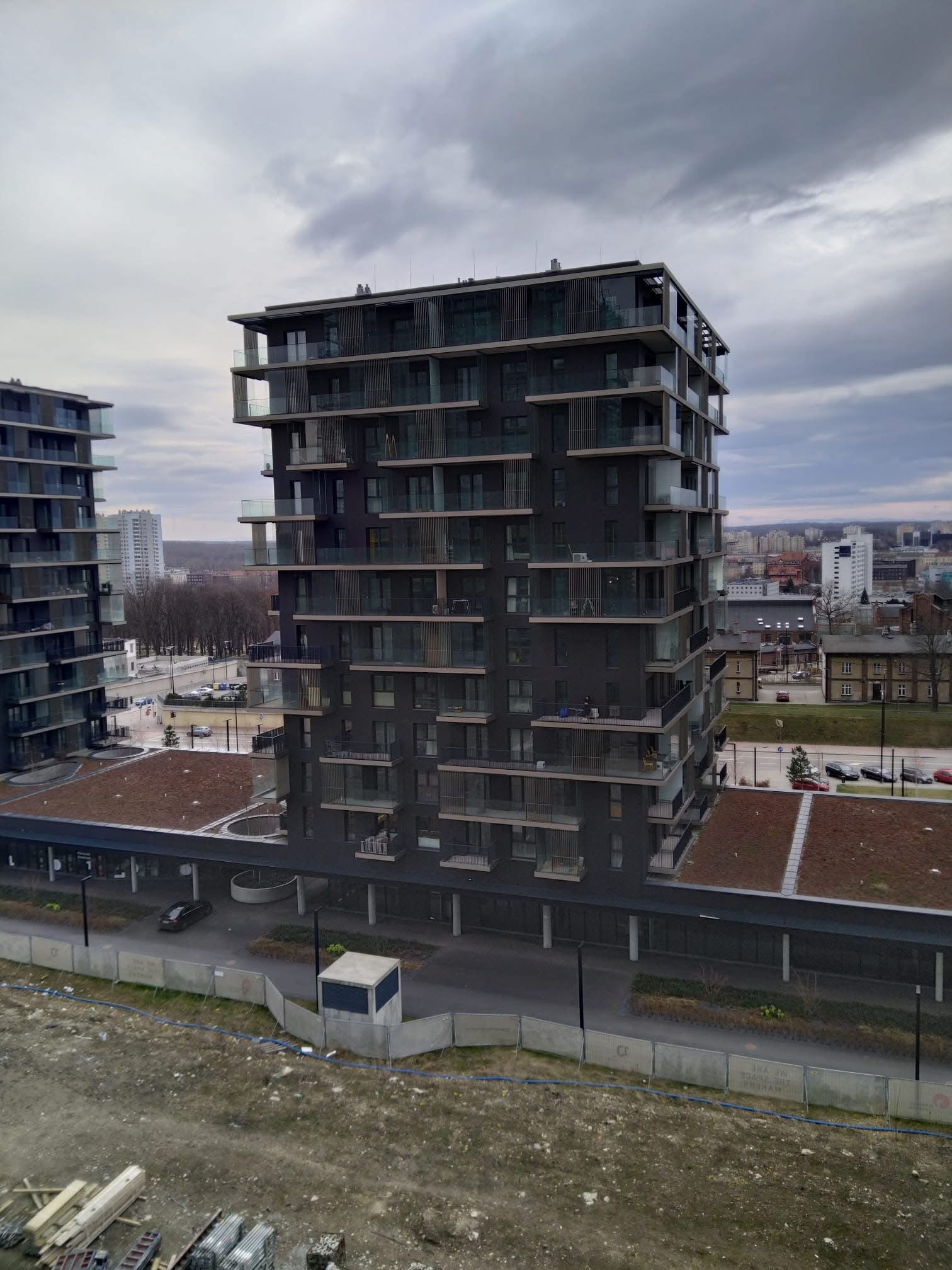 Nowoczesny, wielopiętrowy apartamentowiec z balkonami i zielonym dachem, widoczny z góry na tle pochmurnego nieba i miejskiego krajobrazu. W tle drugi budynek.