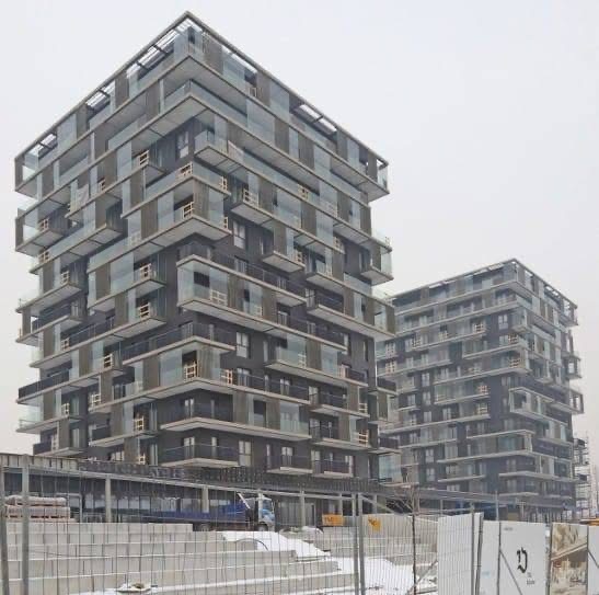 Dwa nowoczesne, wielopiętrowe budynki mieszkalne w Dąbrowie Górniczej, w trakcie budowy. Widoczne szklane balkony i surowa konstrukcja. Ujęcie z dołu, pochmurne niebo.