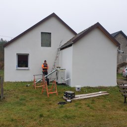 Term - Dach Usługi Elewacyjno-Dekarskie Kamil Dymek - Biała elewacja domu po tynkowaniu, z widocznymi narzędziami i pracownikiem w odblaskowej kamizelce przy drabinie. Świeżo otynkowana ściana z oknem.