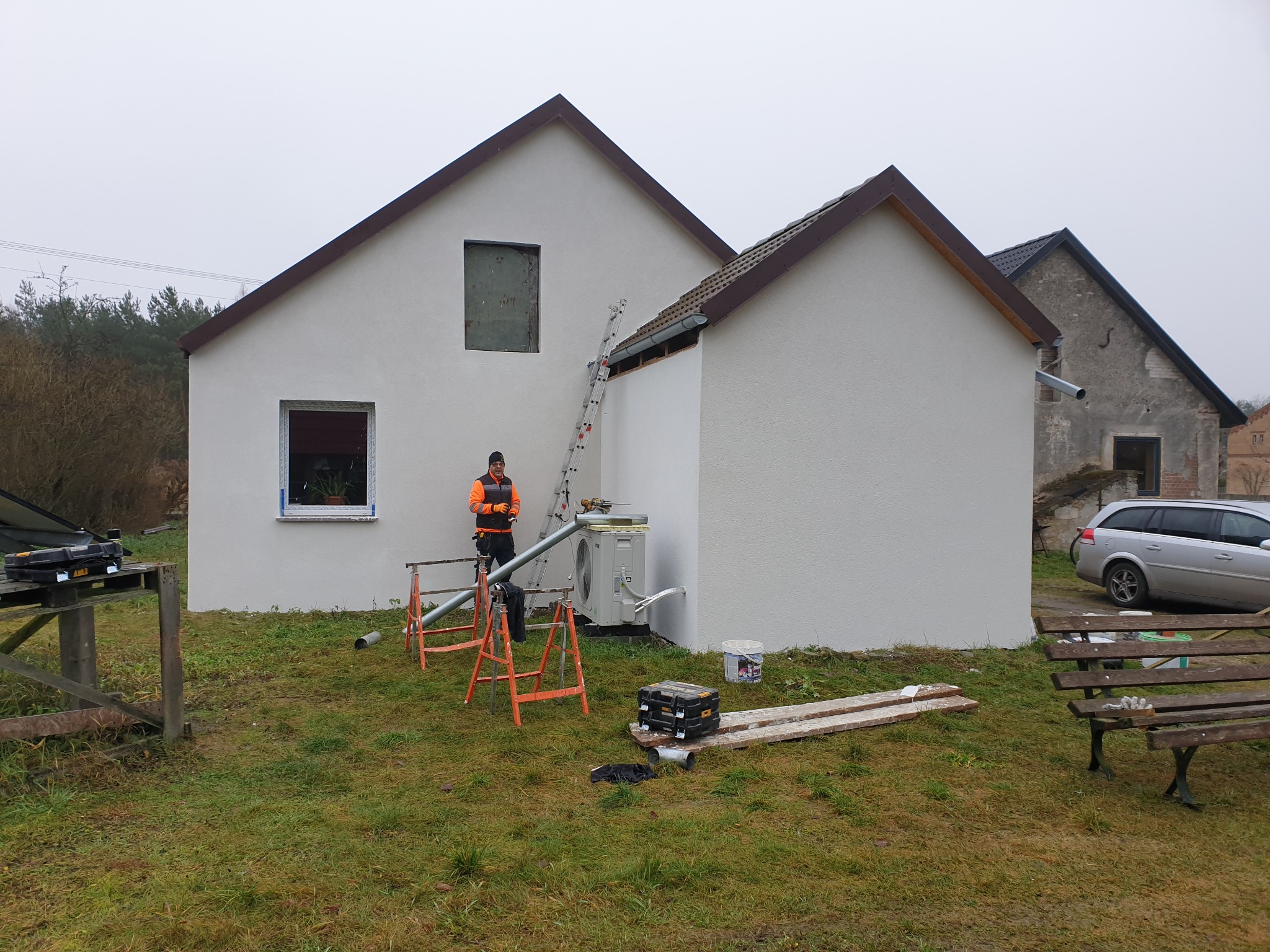 Biała elewacja domu po tynkowaniu, z widocznymi narzędziami i pracownikiem w odblaskowej kamizelce przy drabinie. Świeżo otynkowana ściana z oknem.