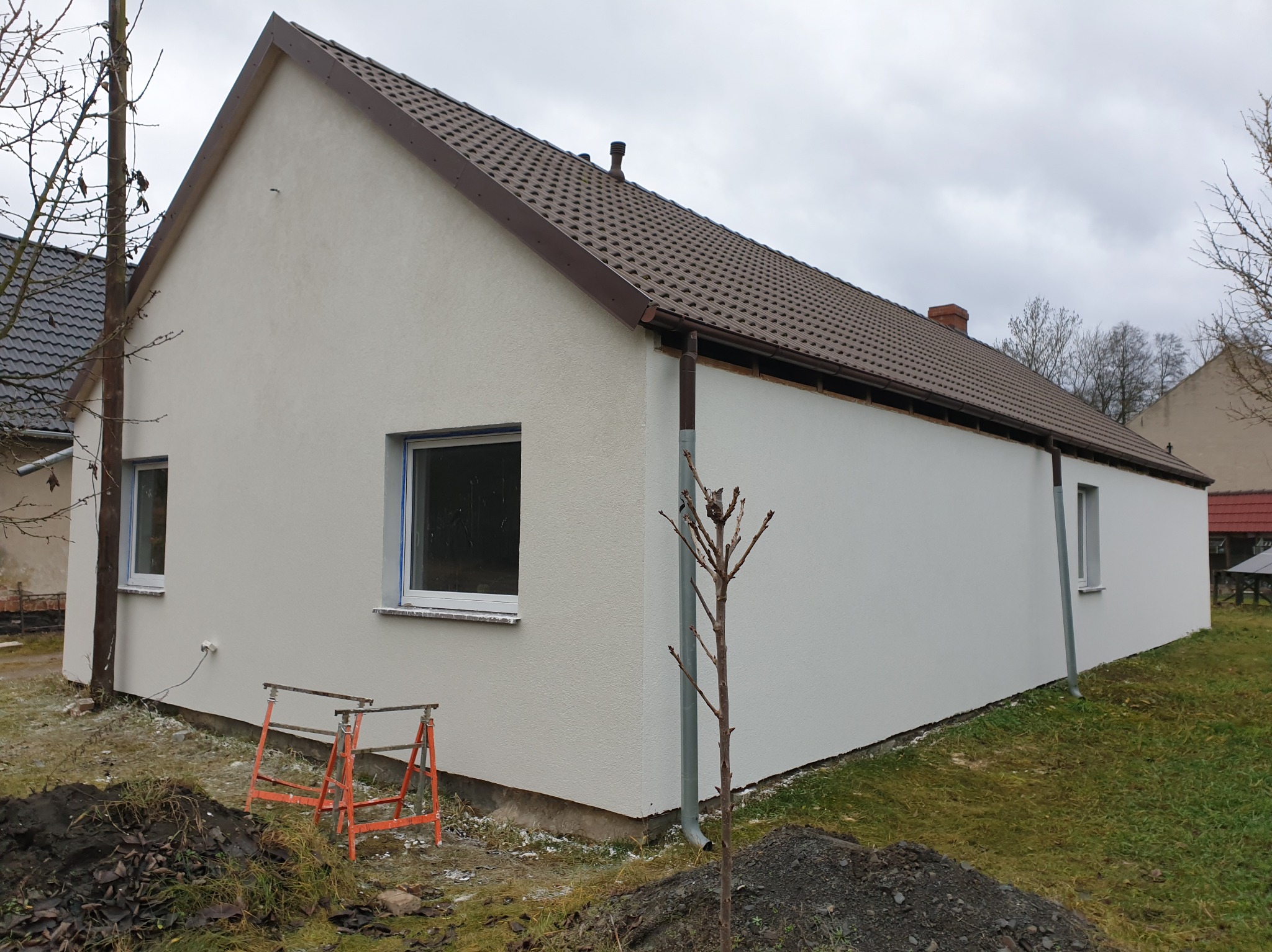 Jasny, parterowy dom z brązowym dachem po tynkowaniu elewacji. Widoczne rusztowania malarskie i fragmenty ogrodu. Pochmurne niebo w tle.