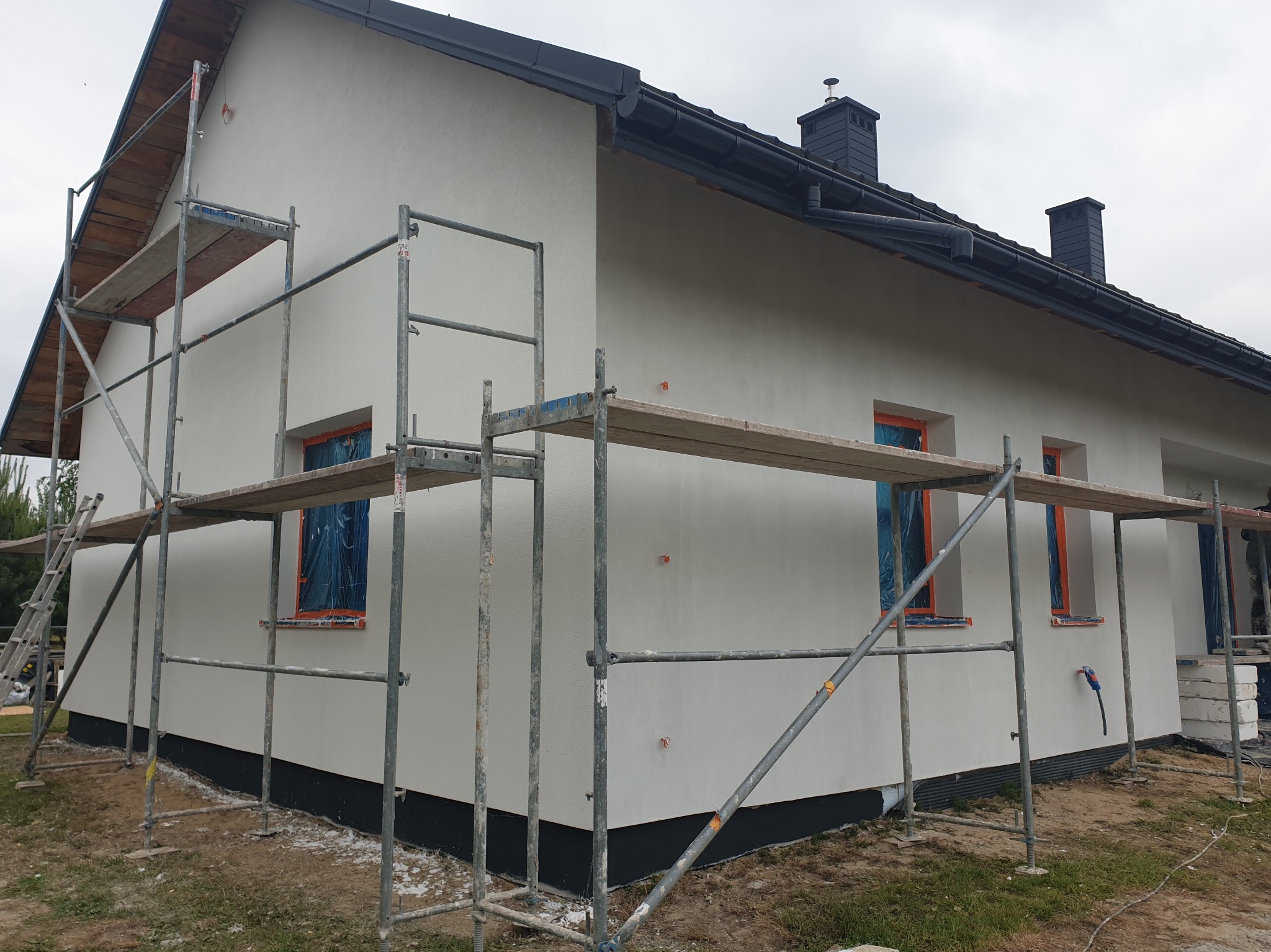 Świeżo otynkowany dom jednorodzinny w jasnym kolorze, z widocznymi rusztowaniami i zabezpieczonymi oknami folią. Nowoczesna bryła budynku z ciemną podmurówką i dachem.
