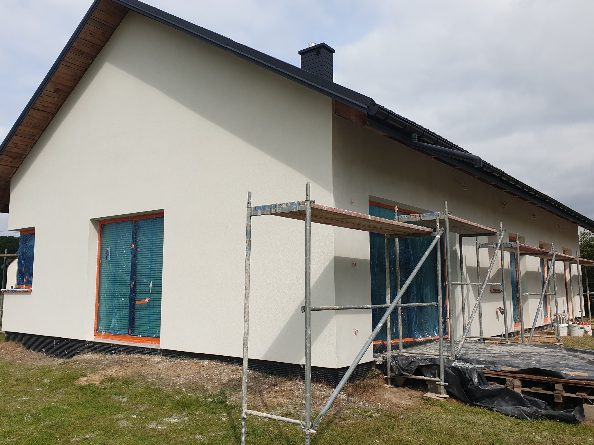 Dom jednorodzinny po tynkowaniu w Trzciance, z oknami zabezpieczonymi folią i rusztowaniem. Widoczny fragment dachu i komin.