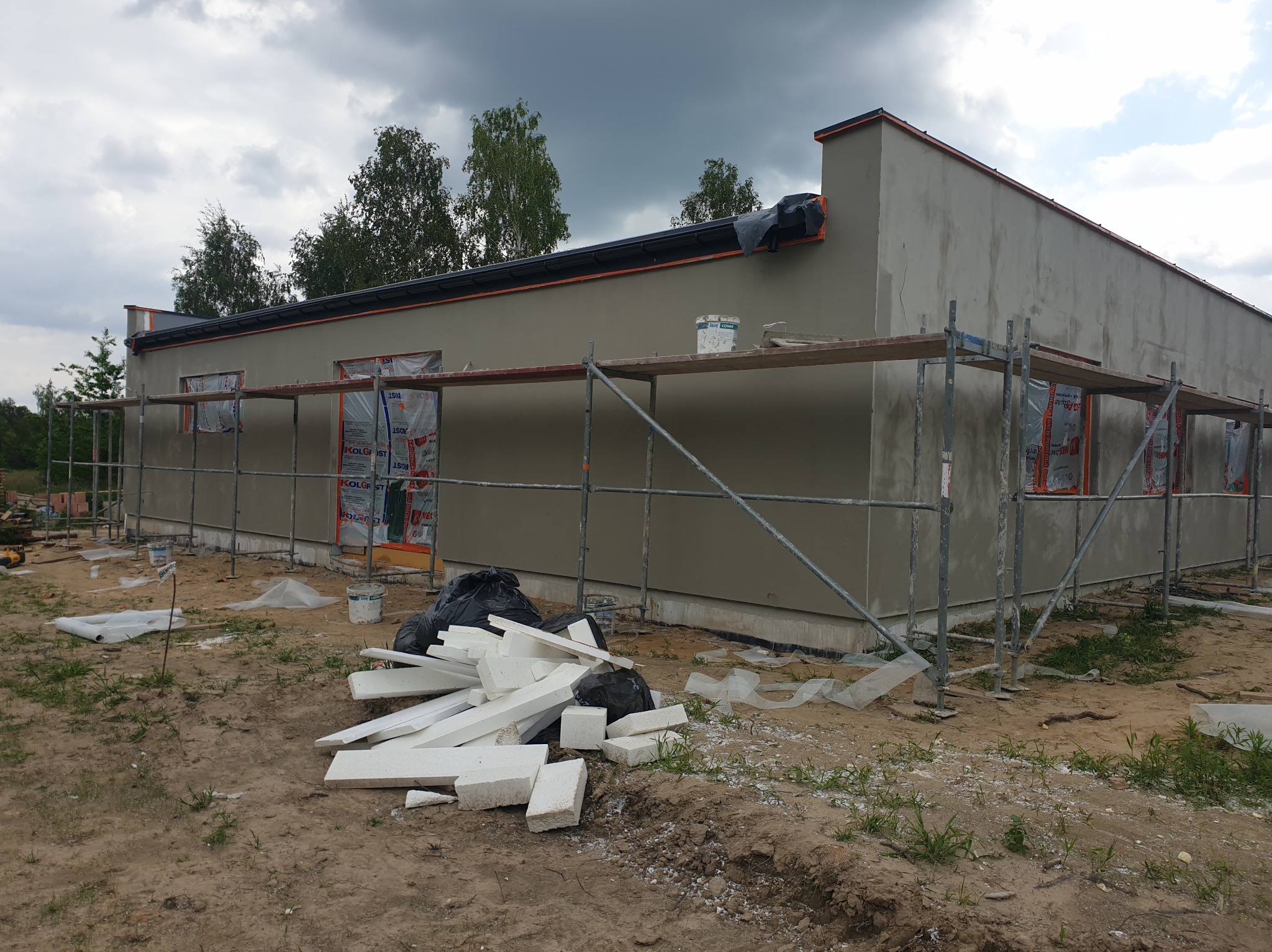 Budynek w trakcie tynkowania elewacji, widoczne rusztowania, styropian i wiadro z zaprawą. Szara elewacja kontrastuje z zielenią drzew i pochmurnym niebem.