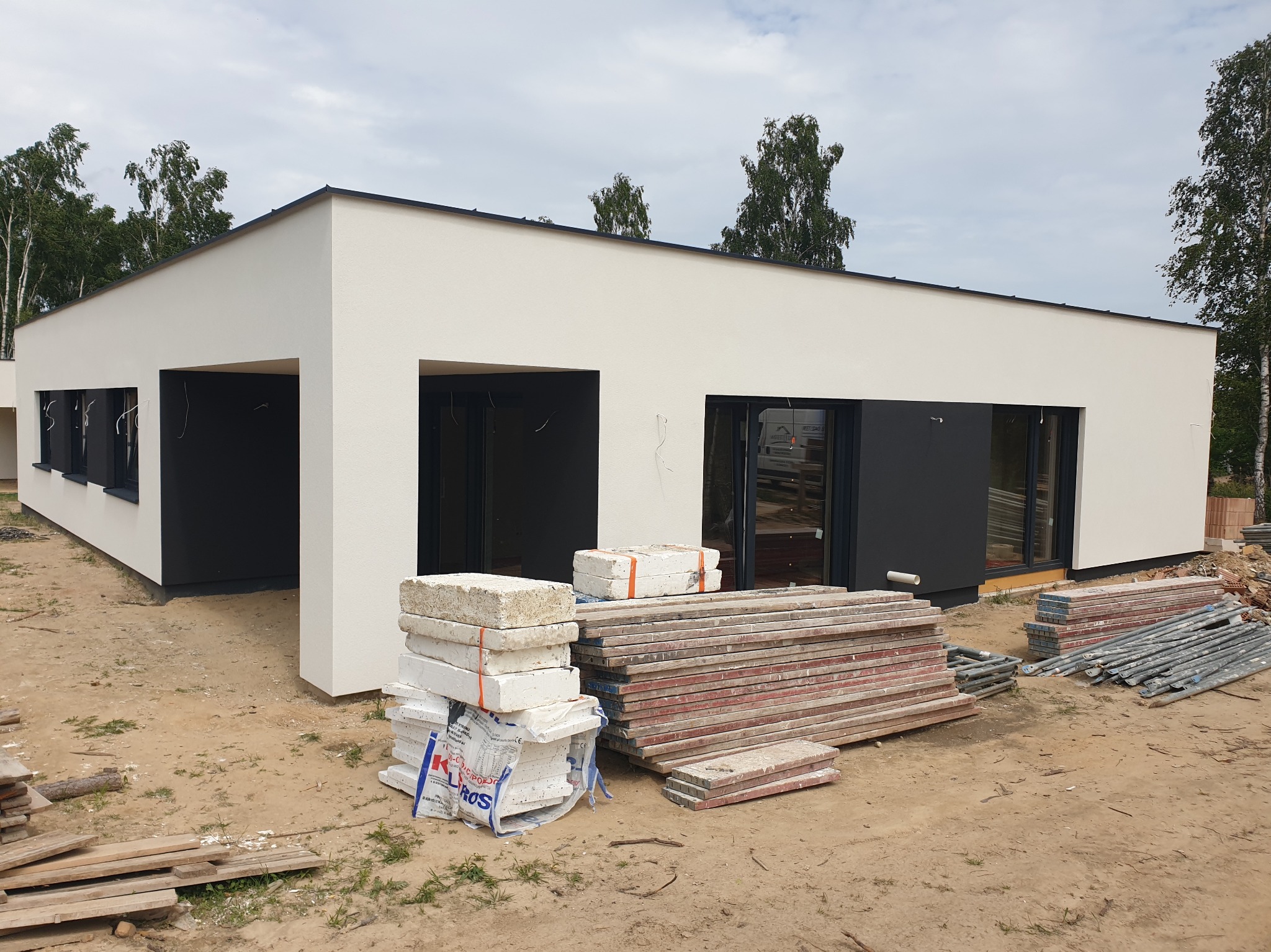 Nowoczesny, parterowy dom z płaskim dachem po tynkowaniu elewacji. Widoczne materiały budowlane i rusztowania. Minimalistyczny design, jasna elewacja, ciemne okna.