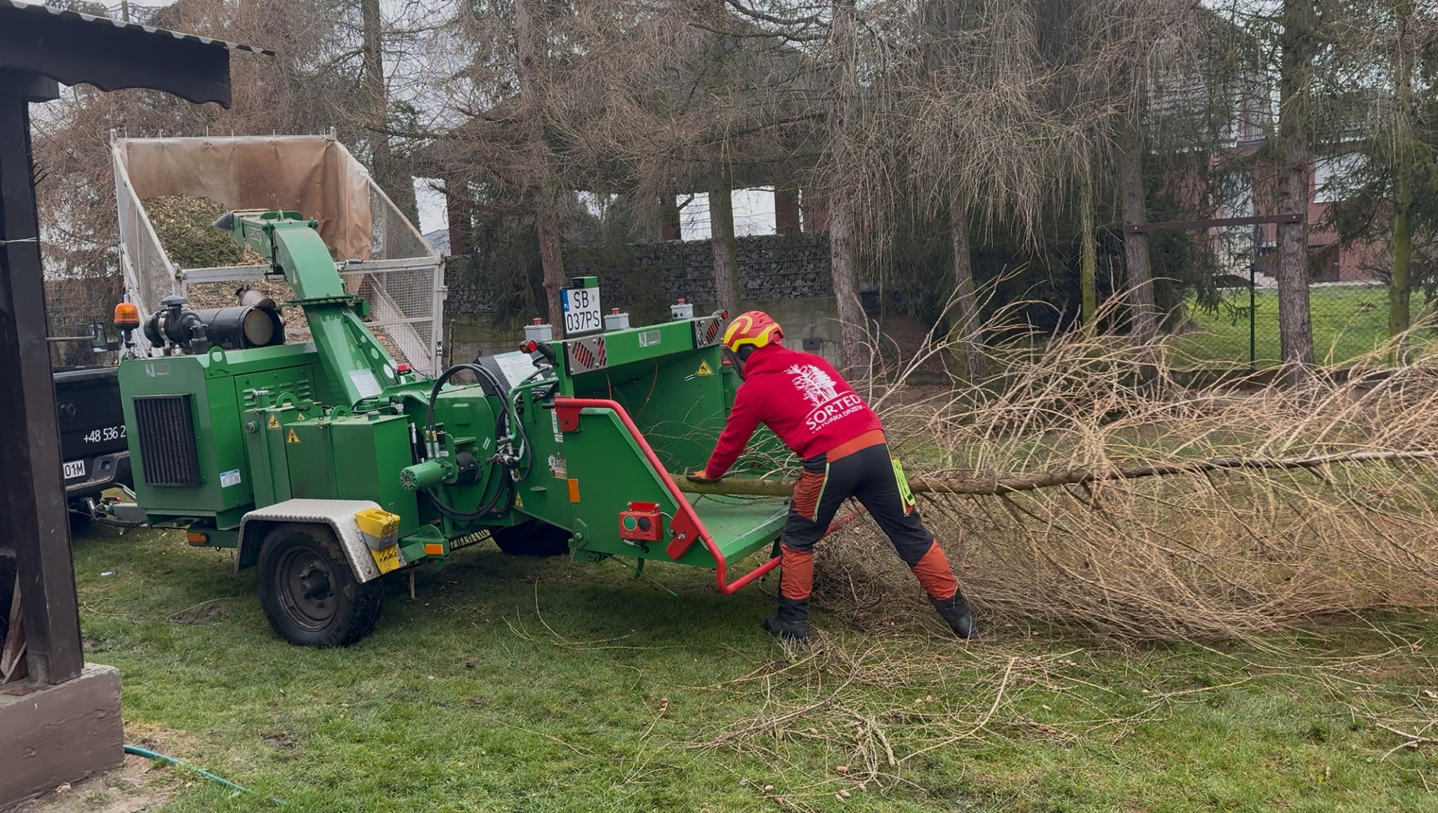 Pracownik w kasku wrzuca gałęzie do zielonego rębaka, który stoi na trawie. W tle budynek i drzewa bez liści. Widoczny fragment przyczepy i tablica rejestracyjna.