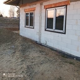 DASIE Usługi Remontowe Dawid Siewiera - Nowe okna PCV z brązowymi ramami zamontowane w surowym budynku z białych bloczków. Widoczny piasek i fragment krajobrazu w odbiciu szyb.