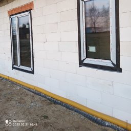 DASIE Usługi Remontowe Dawid Siewiera - Dwa okna PCV z czarnymi ramami zamontowane w ścianie z białych bloczków. Żółta rura drenażowa wzdłuż fundamentu. Widok z zewnątrz, etap budowy.