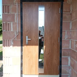 DASIE Usługi Remontowe Dawid Siewiera - Zamontowane drzwi wejściowe z przeszkleniem, widoczne na tle surowej ściany z cegły. Na drzwiach widoczne oznaczenia producenta. W odbiciu szyby widać osobę robiącą zdjęcie.