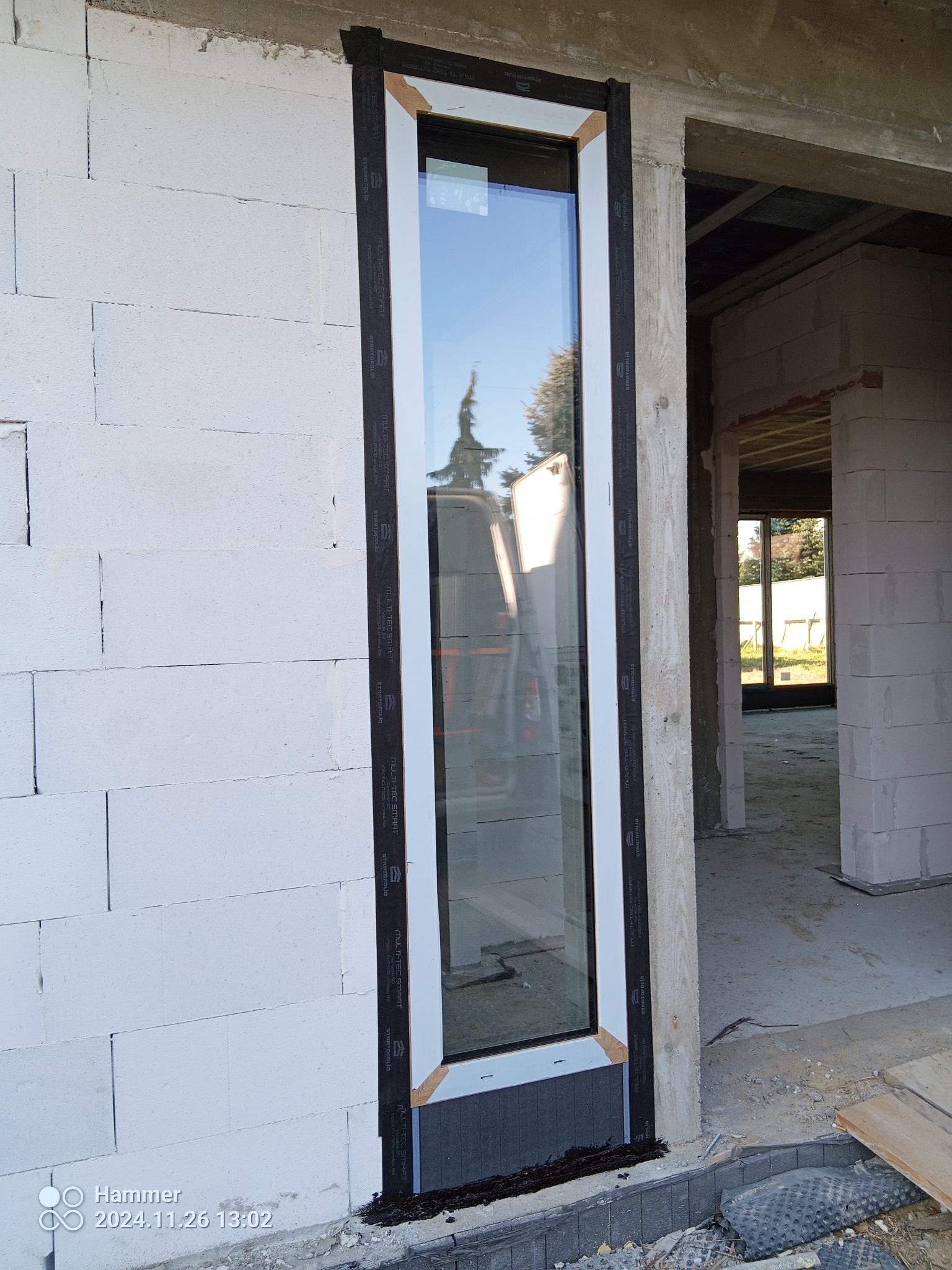 Wąskie okno PCV w surowym murze z betonu komórkowego, zabezpieczone taśmą montażową. Widok na wnętrze budynku w stanie deweloperskim i odbicie drzewa w szybie.