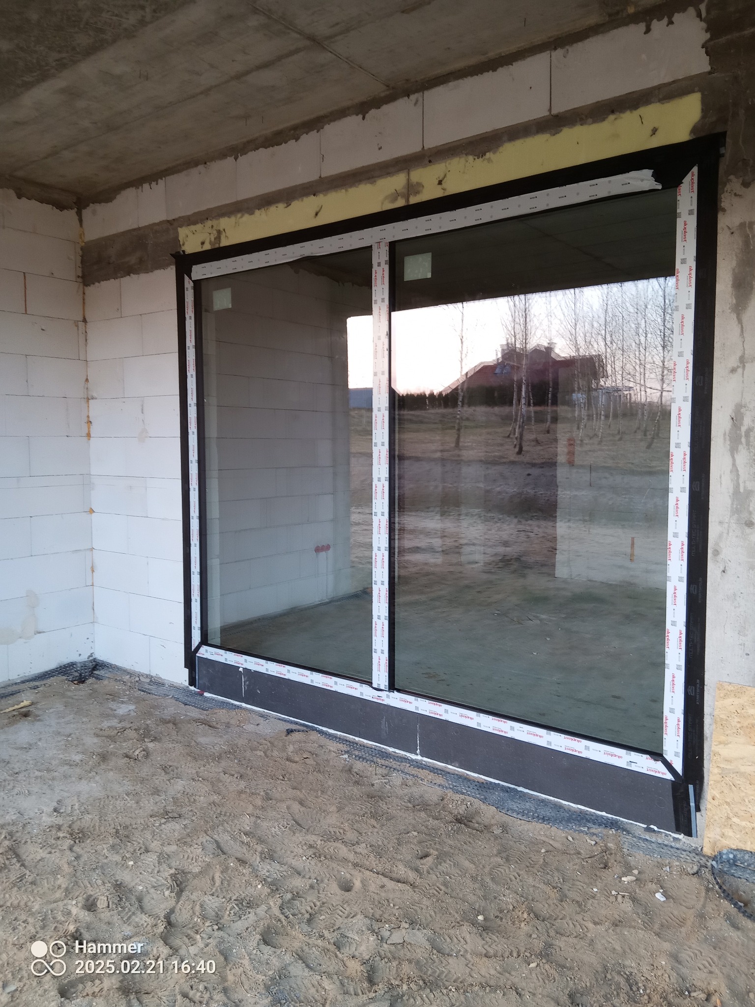 Montaż okna PCV z czarną ramą w surowym stanie budynku w Rzeszowie. Widoczne bloczki betonowe i piasek na podłodze. Okno marki aluplast.