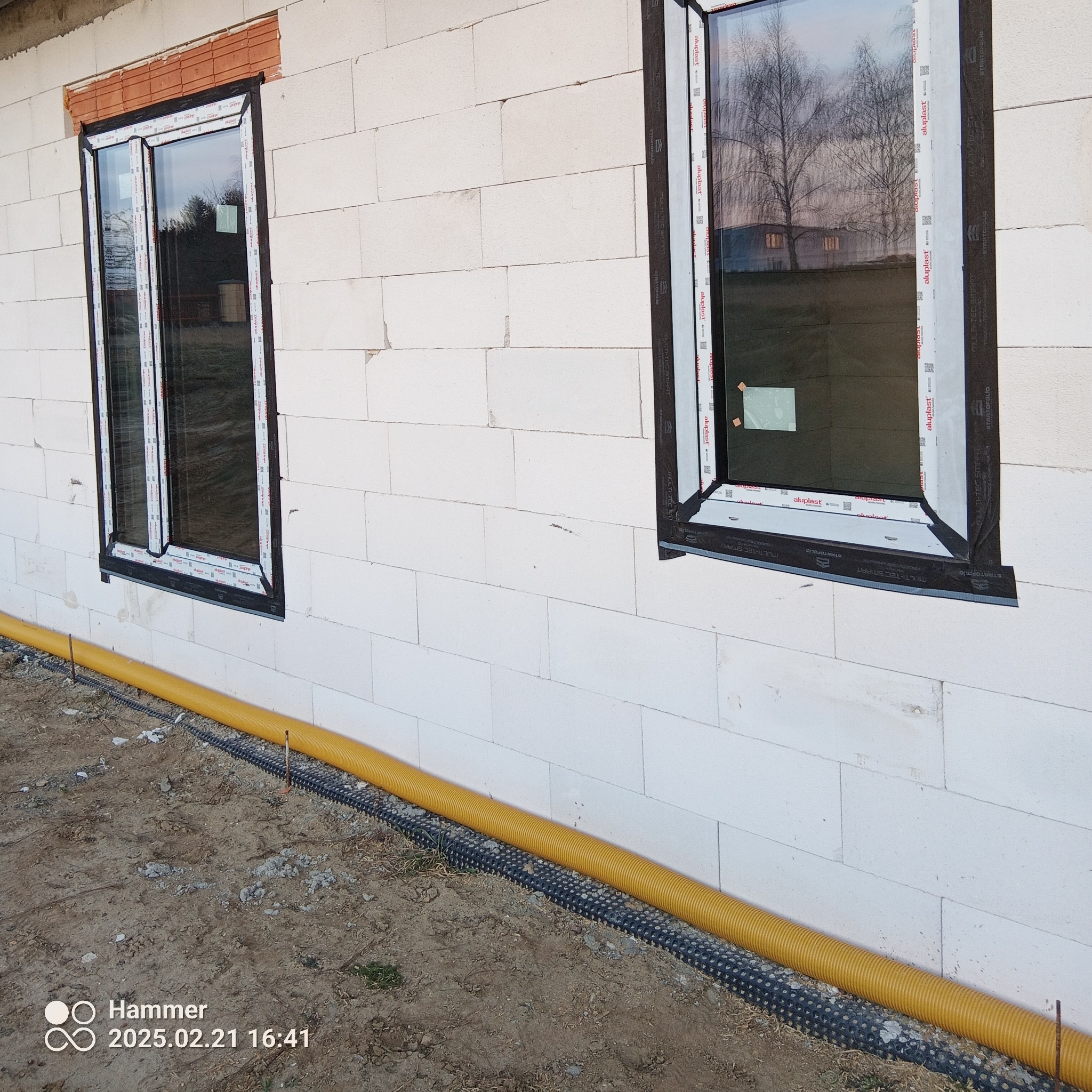 Dwa okna PCV z czarnymi ramami zamontowane w ścianie z białych bloczków. Żółta rura drenażowa wzdłuż fundamentu. Widok z zewnątrz, etap budowy.