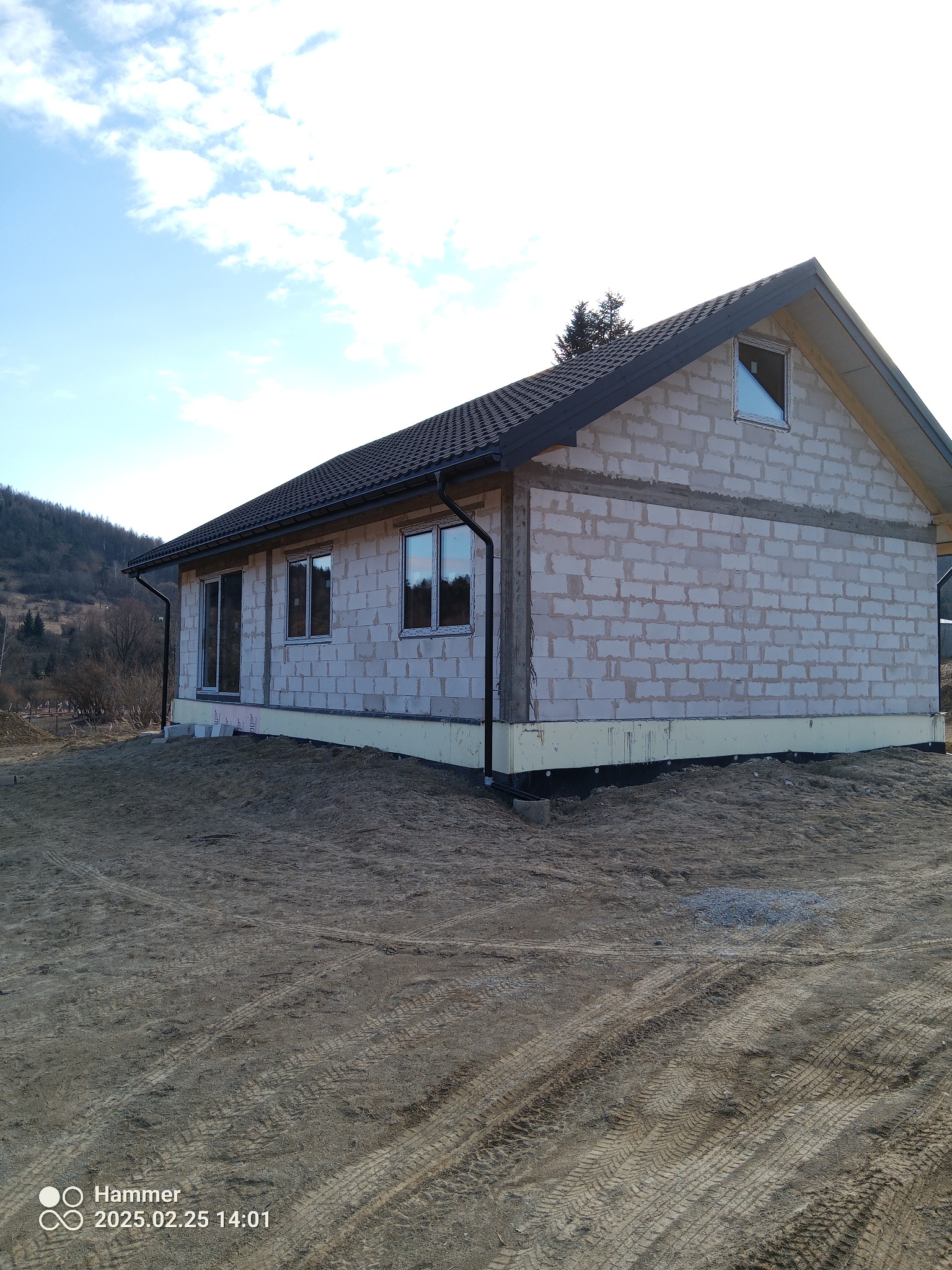 Surowy stan domu jednorodzinnego z białego bloczka, z zamontowanymi oknami PCV i czarną dachówką, na tle wzgórz i błękitnego nieba z chmurami.