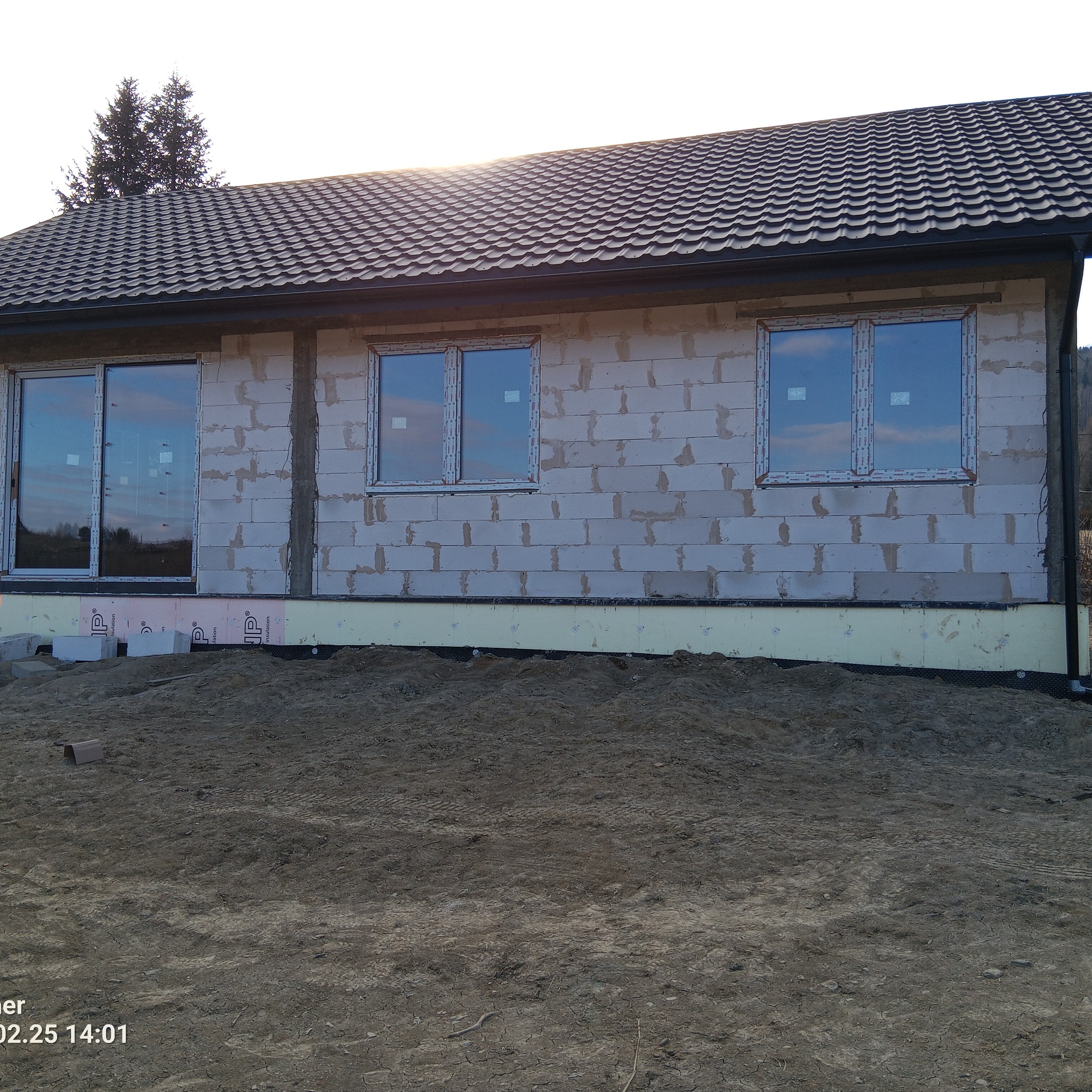 Surowy stan budynku z zamontowanymi oknami PCV, widoczne bloczki betonowe i izolacja fundamentów. W tle drzewa i niebo.