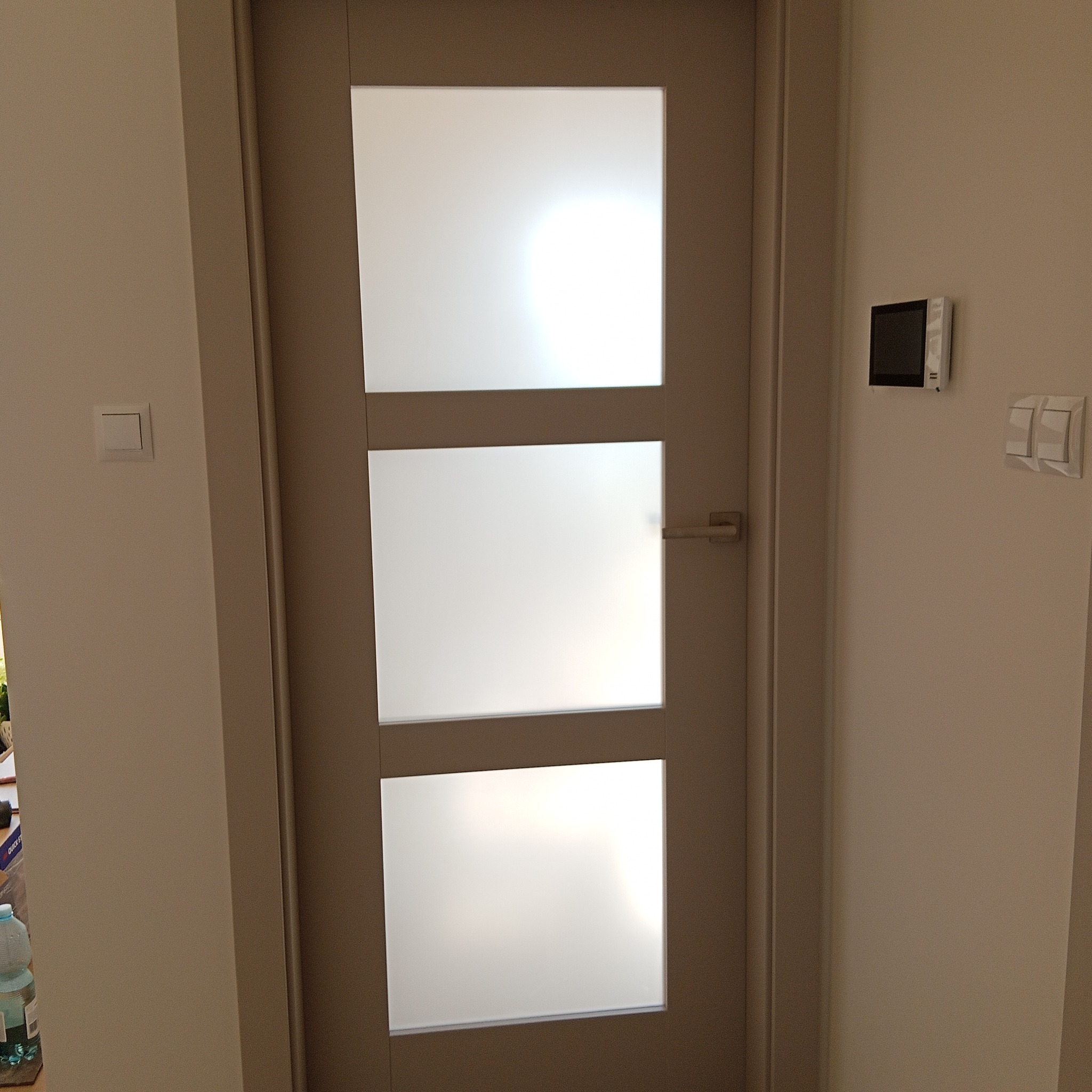 Beżowe drzwi wewnętrzne z trzema matowymi szybami, widoczne na tle jasnej ściany z włącznikami. Minimalistyczny design, proste linie, nowoczesny styl.