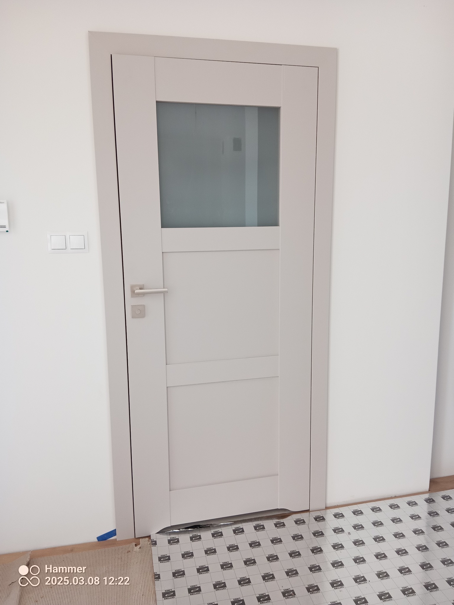 Zamontowane, wewnętrzne drzwi w odcieniach szarości z szybą, widoczne na tle białej ściany i podłogi zabezpieczonej folią ochronną. Minimalistyczny design.