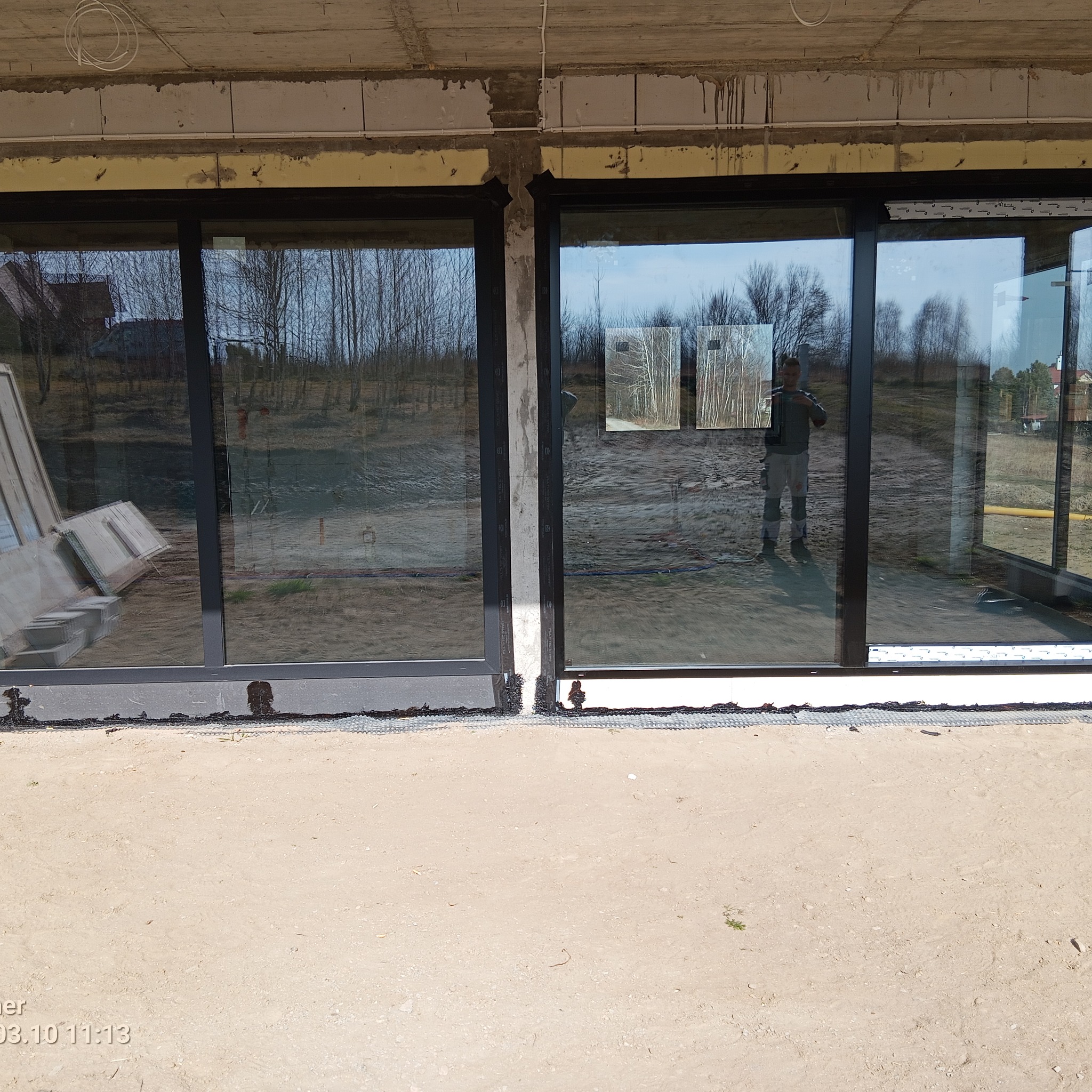 Czarne okna przesuwne zamontowane w surowym stanie budynku. Widoczne detale montażu i odbicie osoby w szybie. Na dole piasek.