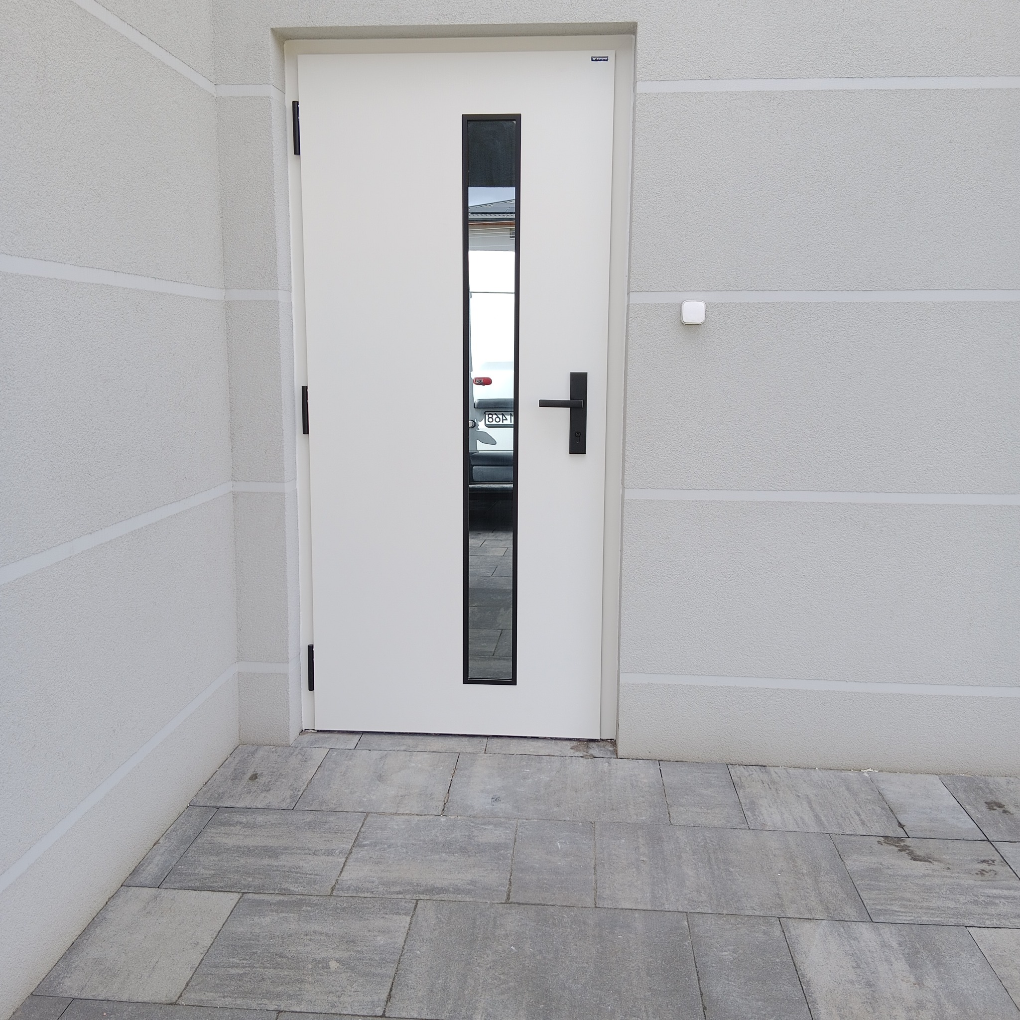 Białe drzwi wejściowe z czarną szybą i klamką, zamontowane w jasnej elewacji z poziomymi pasami. Widoczna szara kostka brukowa i odbicie samochodu w szybie.