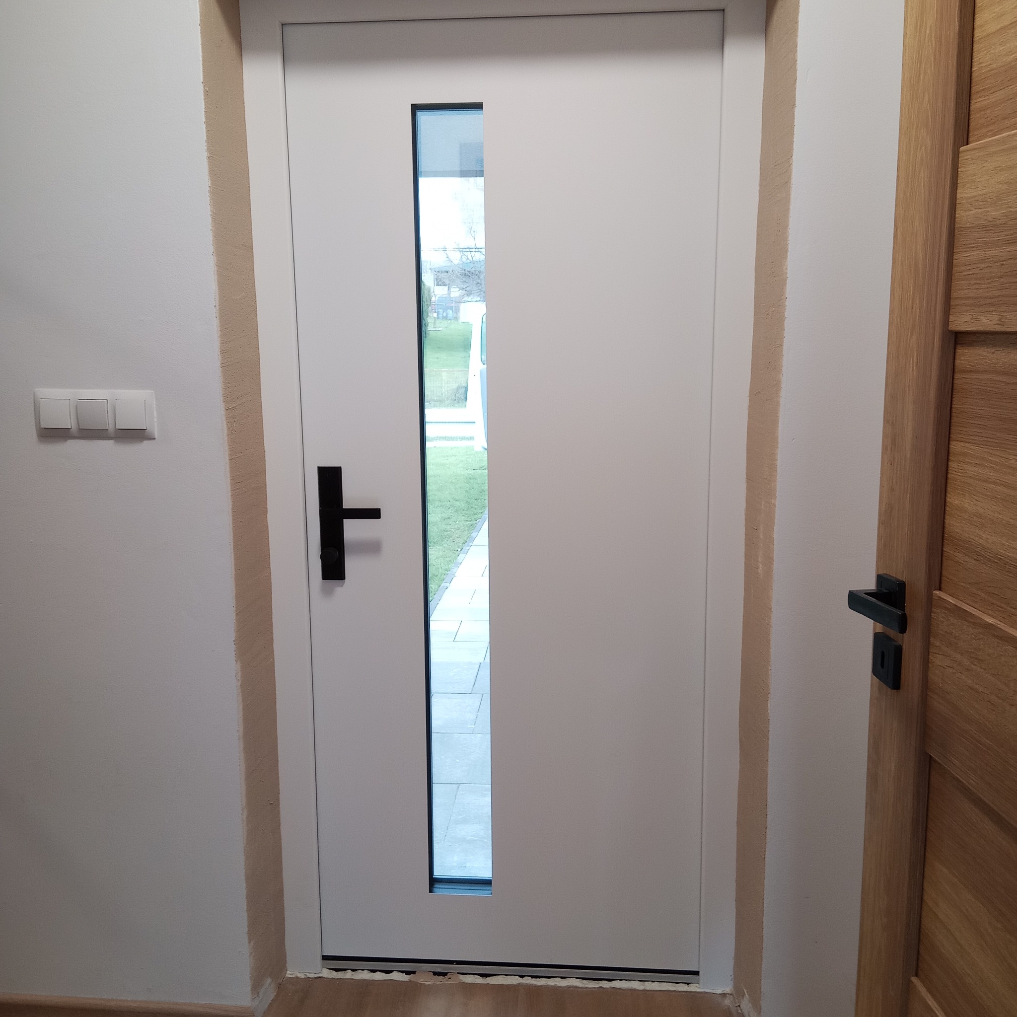 Białe drzwi wejściowe z czarną klamką i wąskim przeszkleniem, przez które widać fragment podwórka. Obok widoczna drewniana ościeżnica innych drzwi.