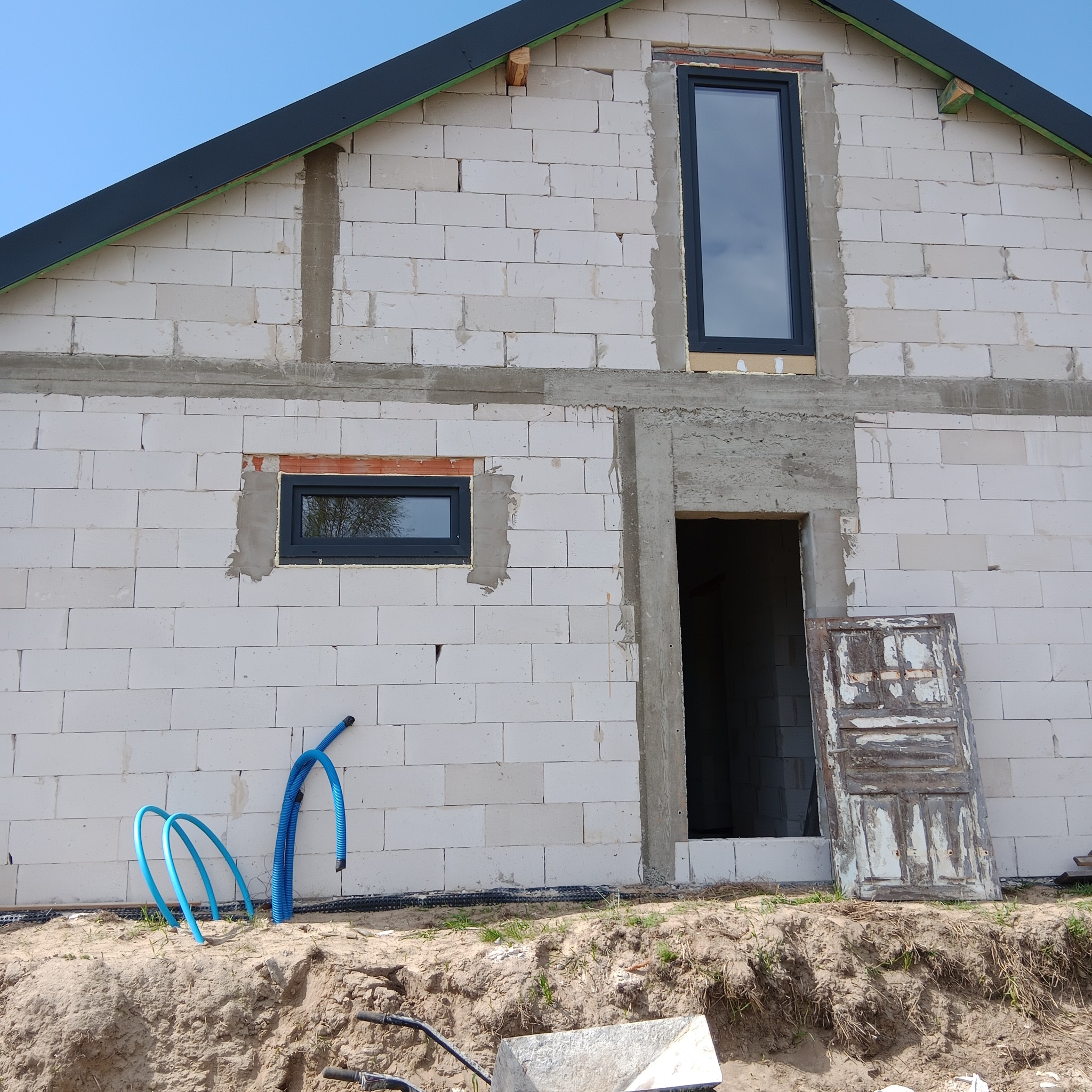 Budowa domu z pustaków: wstawione okna PCV, otwór drzwiowy, niebieskie rury na ziemi, stara, drewniana płycina drzwiowa oparta o ścianę. Widok na budynek w trakcie budowy.