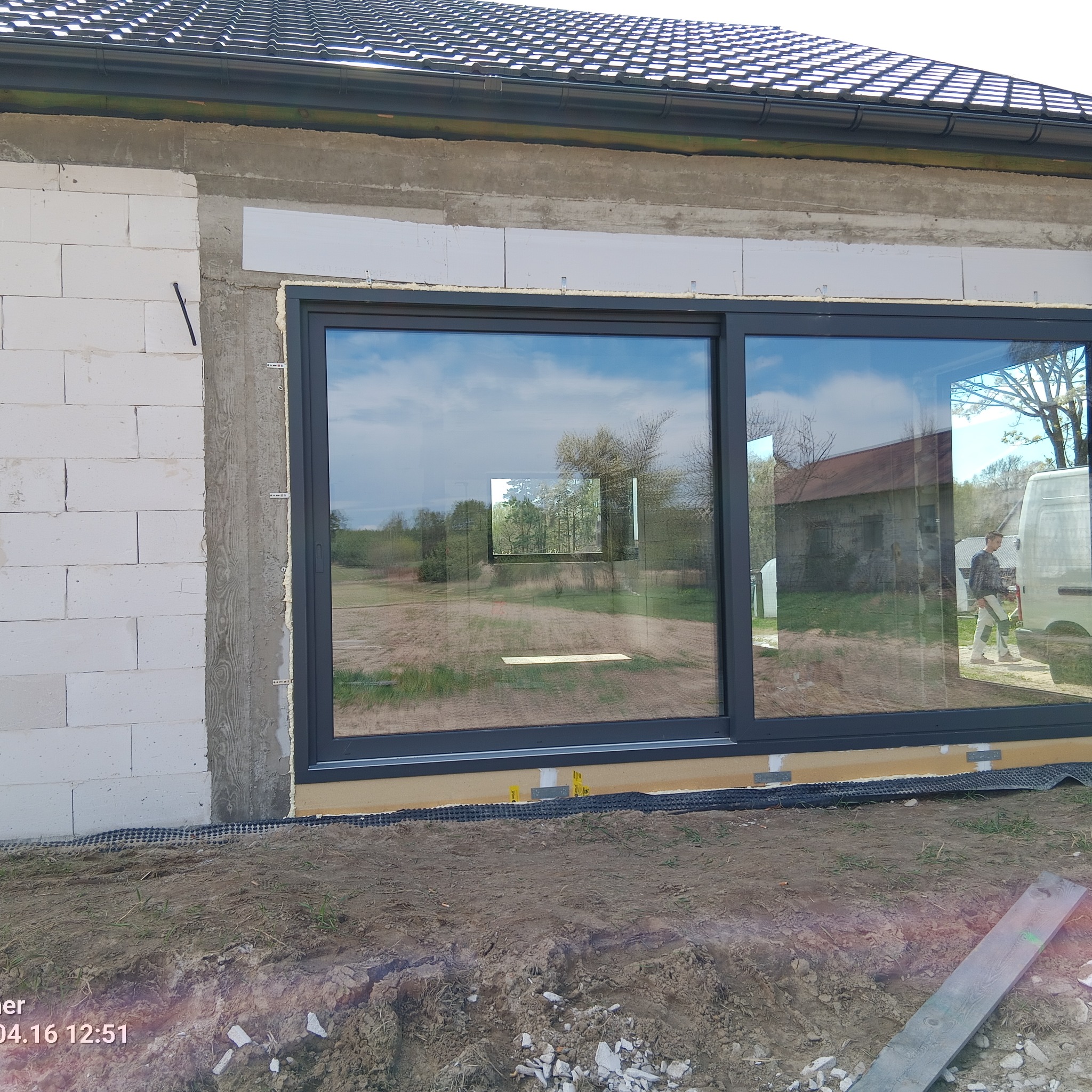 Duże, grafitowe okno przesuwne w surowym murze z betonu komórkowego. Widoczne elementy montażowe i odbicie krajobrazu w szybie. Prace wykończeniowe w toku.