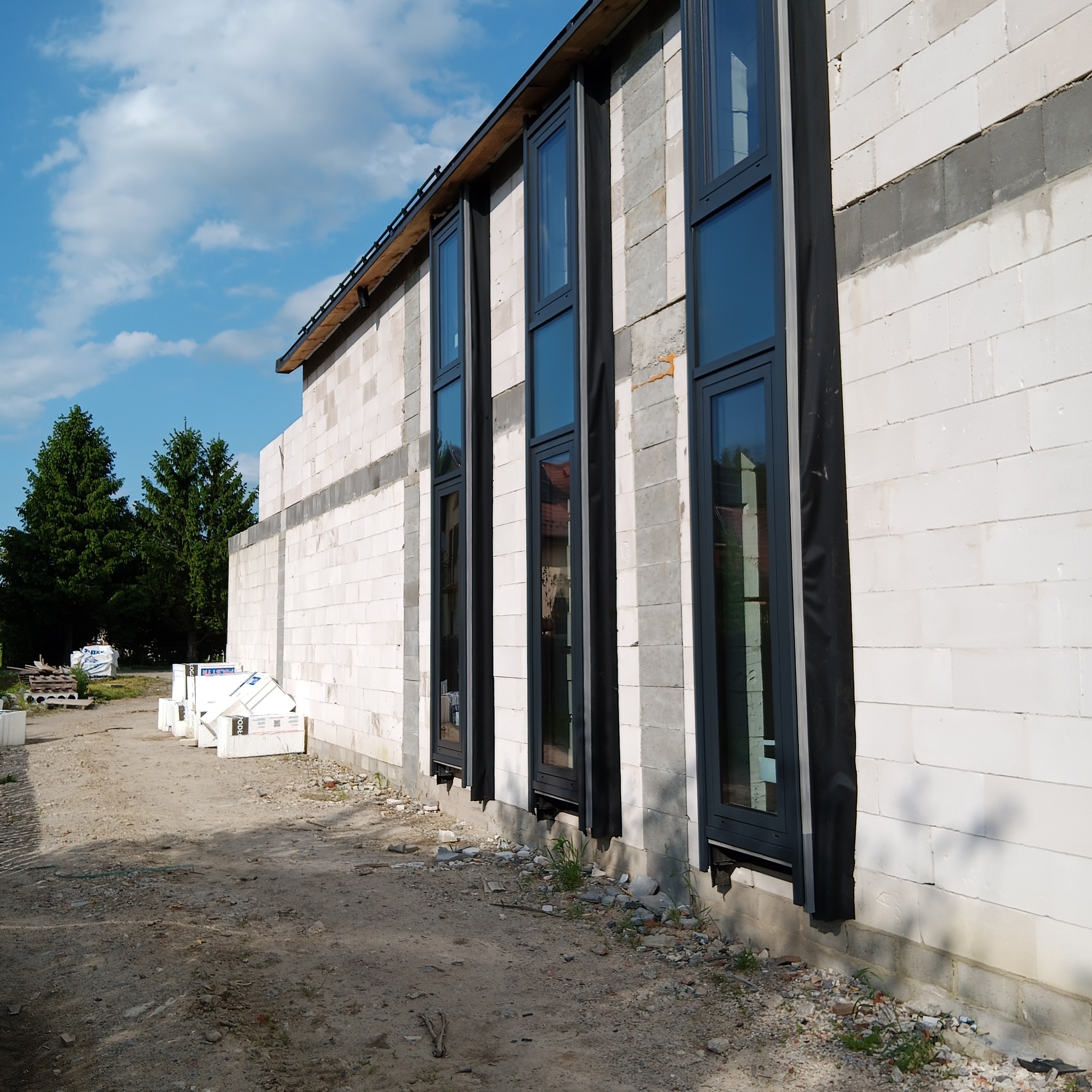 Montaż okien w surowym budynku z betonu komórkowego. Widoczne czarne ramy okienne i zabezpieczenia. Na pierwszym planie gruz i materiały budowlane.