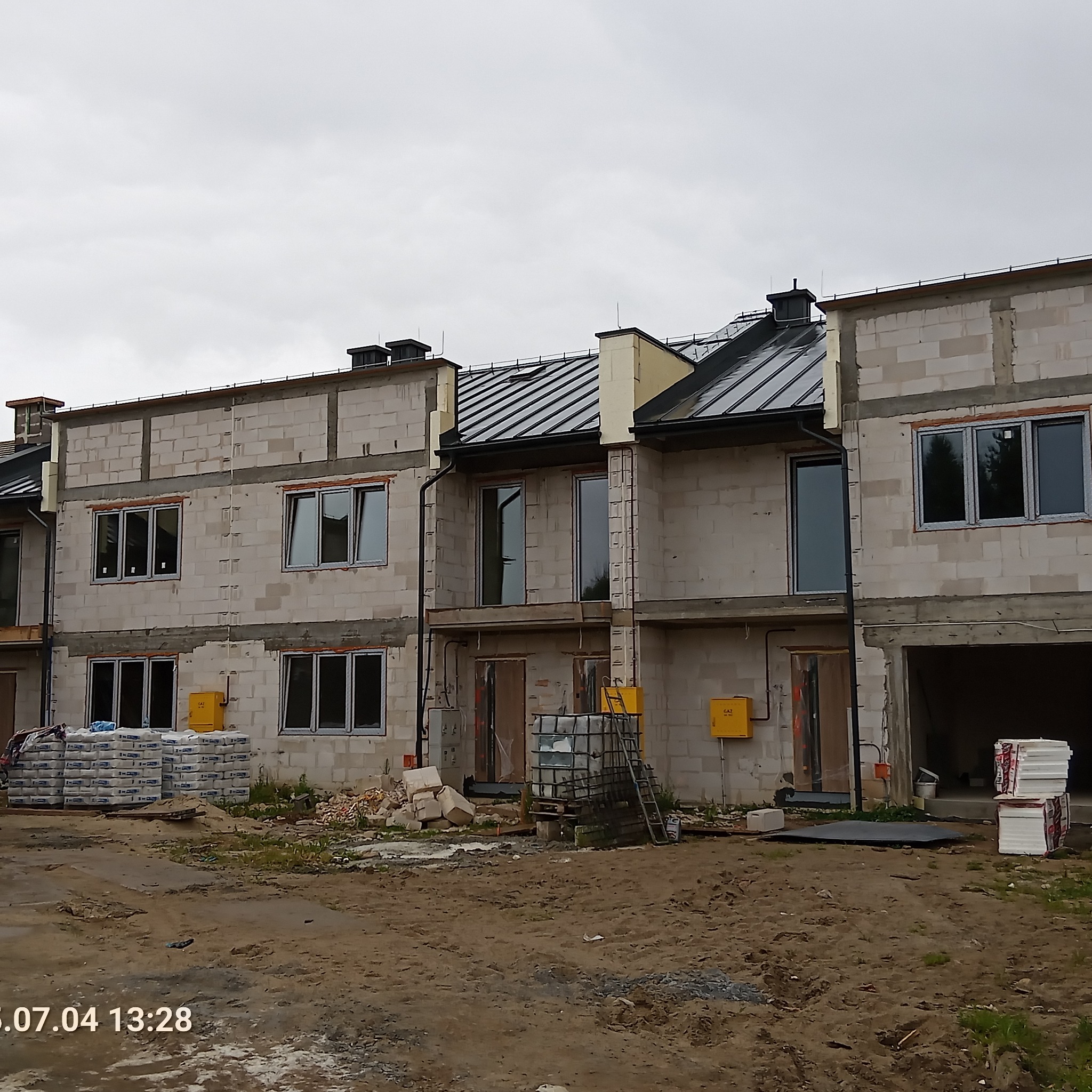 Surowy stan budowy szeregowców z oknami, widoczne materiały budowlane i instalacje. Szare niebo w tle. Ujęcie z dołu, podkreślające skalę projektu.