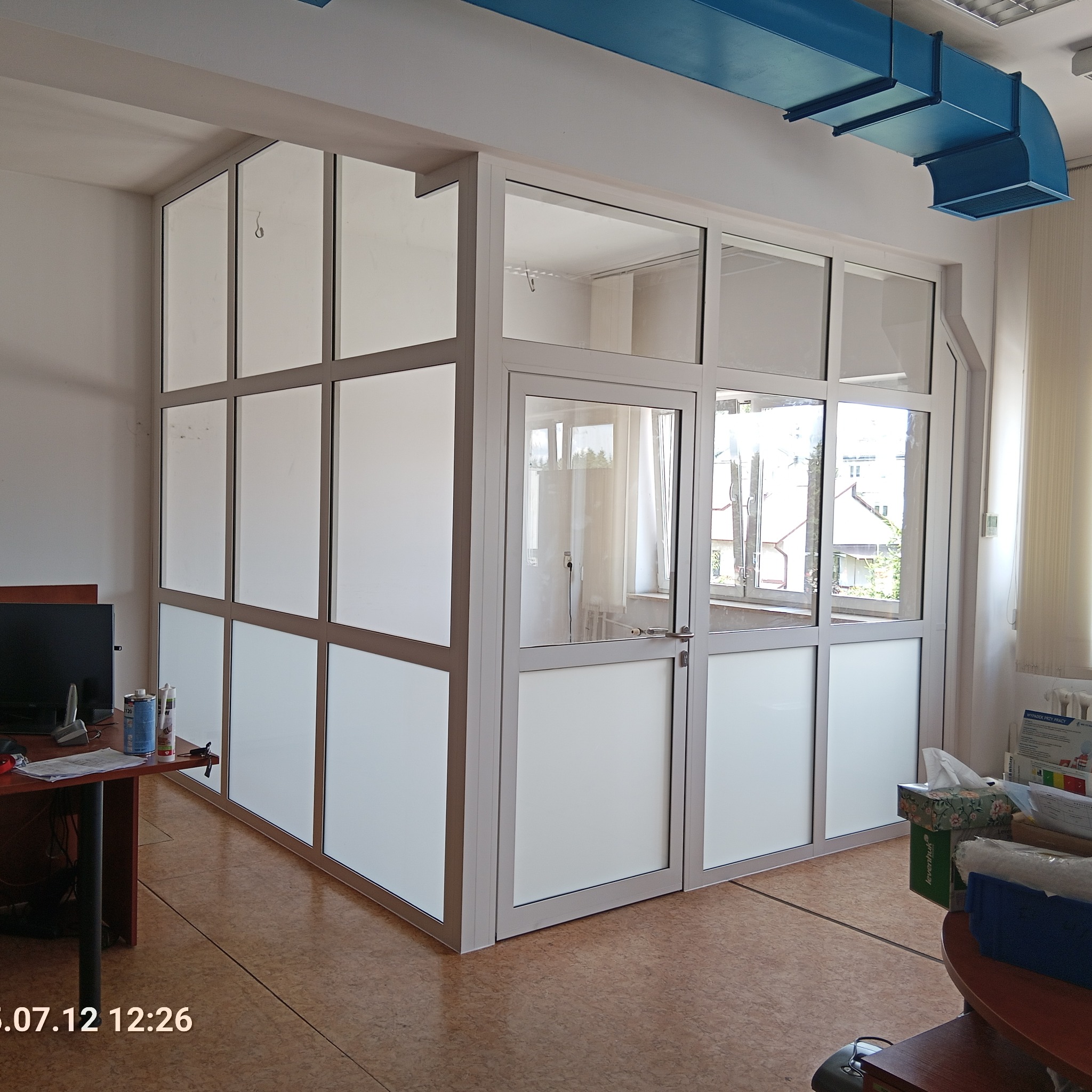 Biała ścianka działowa z drzwiami, wykonana z profili aluminiowych i matowego szkła, oddzielająca przestrzeń biurową. Widoczne biurko i elementy instalacji wentylacyjnej.