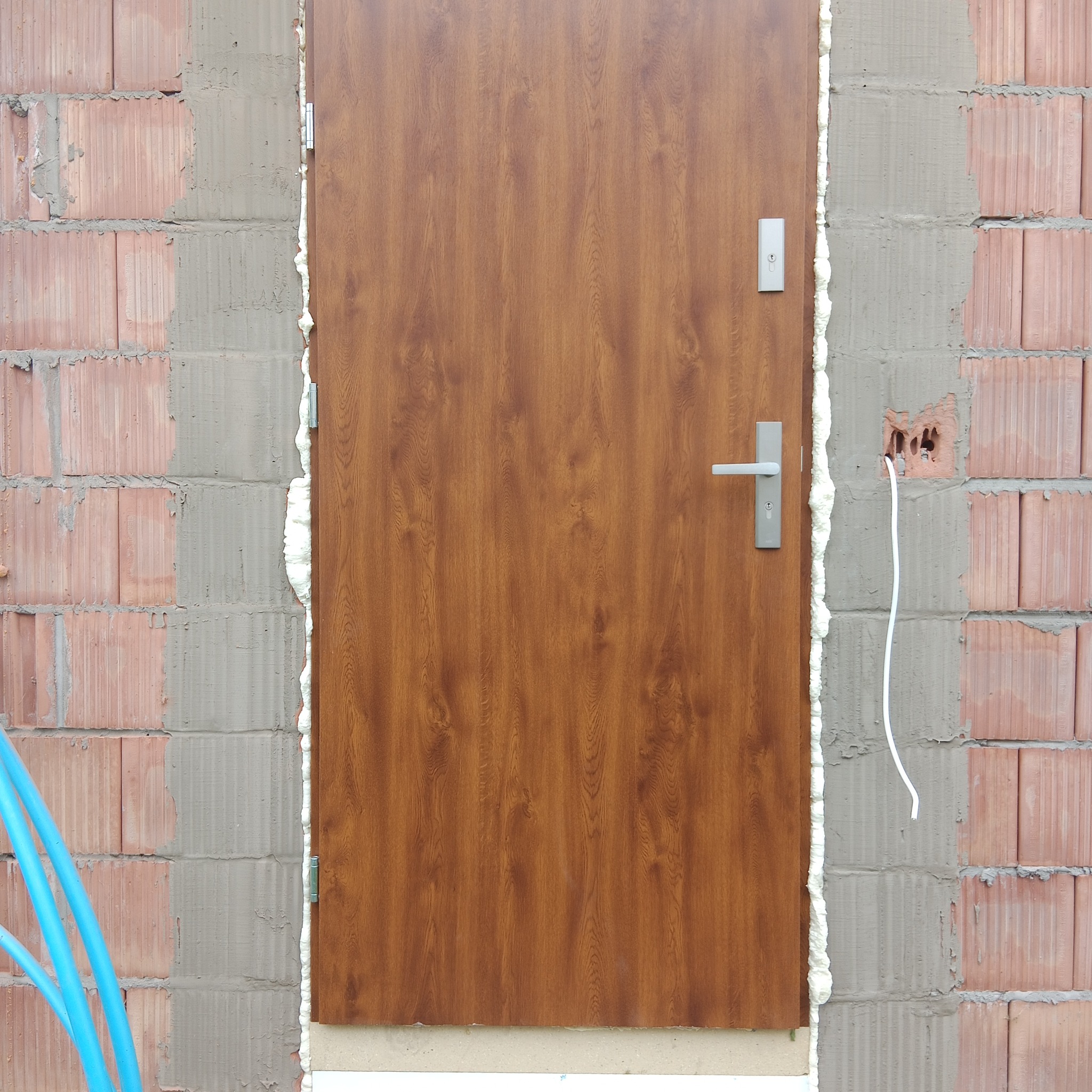 Brązowe drzwi wejściowe z ościeżnicą uszczelnioną pianką montażową, osadzone w surowej ścianie z cegły. Widoczne przewody elektryczne i fragment niebieskich rur.